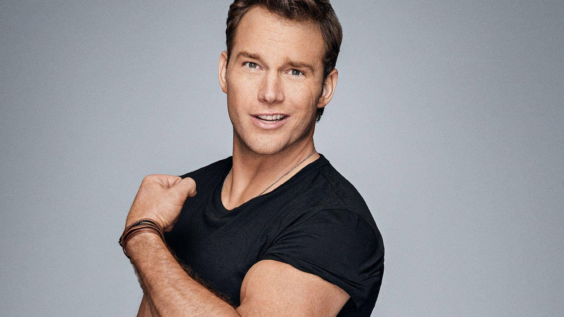 Chris Pratt 桌布 壁紙 — 一位身穿黑色T恤的健壯男子，在純灰色攝影棚背景下自信地微笑並彎曲二頭肌。