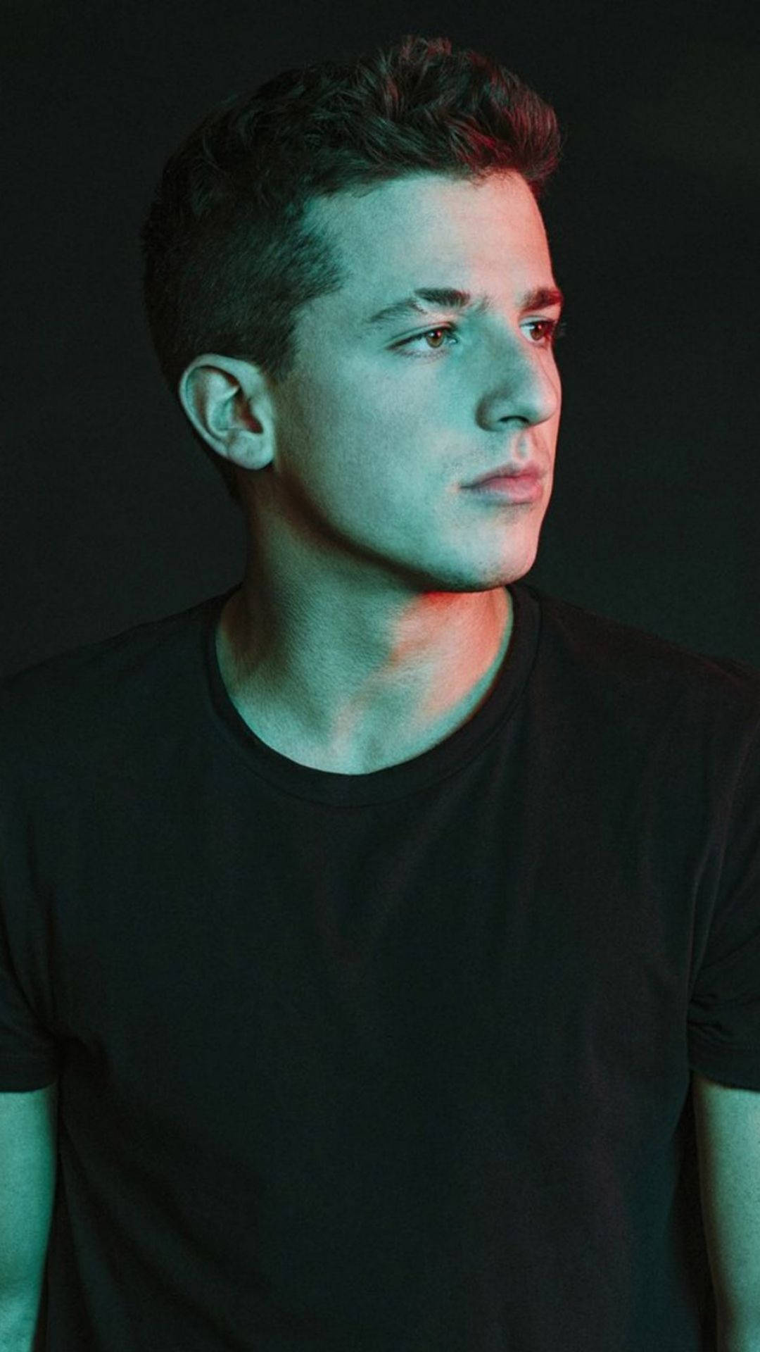Charlie Puth 桌布 壁紙 — 一位身穿黑色T恤的年輕男子，在冷藍色和暖紅色燈光籠罩的黑暗背景下若有所思地凝視著。