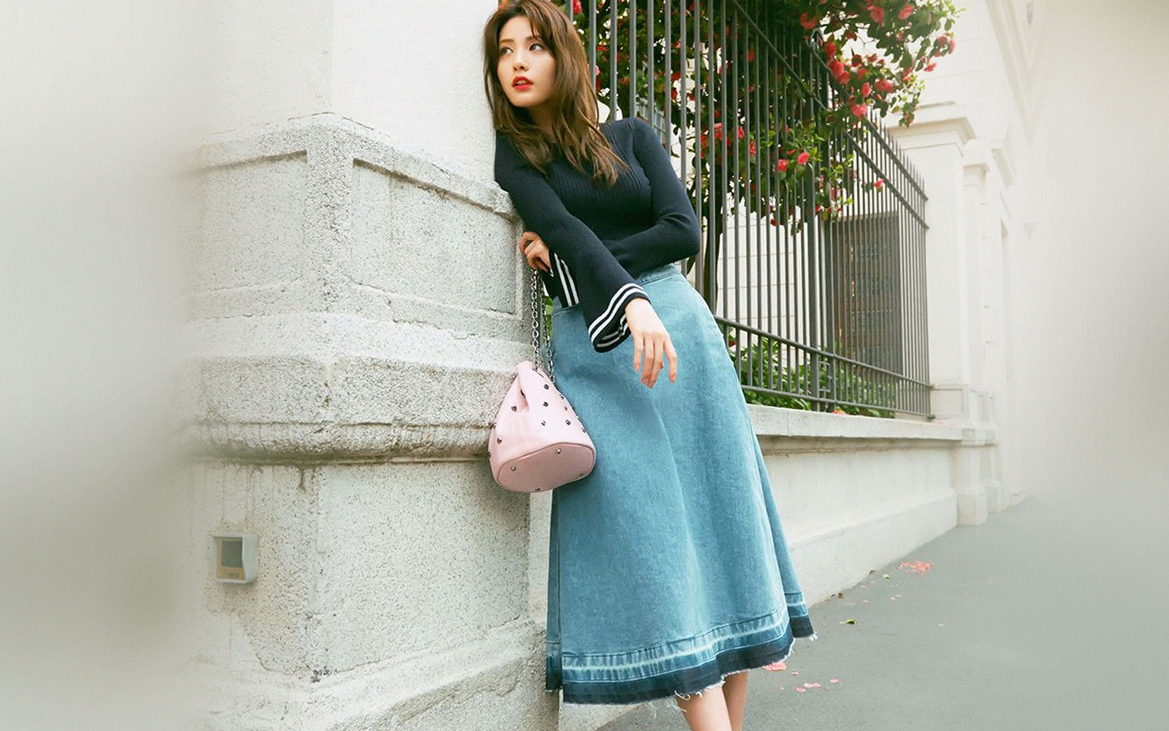 Spring Fashion 桌布 壁紙 — 一位身穿黑色上衣和牛仔裙的女性靠在石牆上，旁邊是帶有紅花的鍛鐵圍欄。
