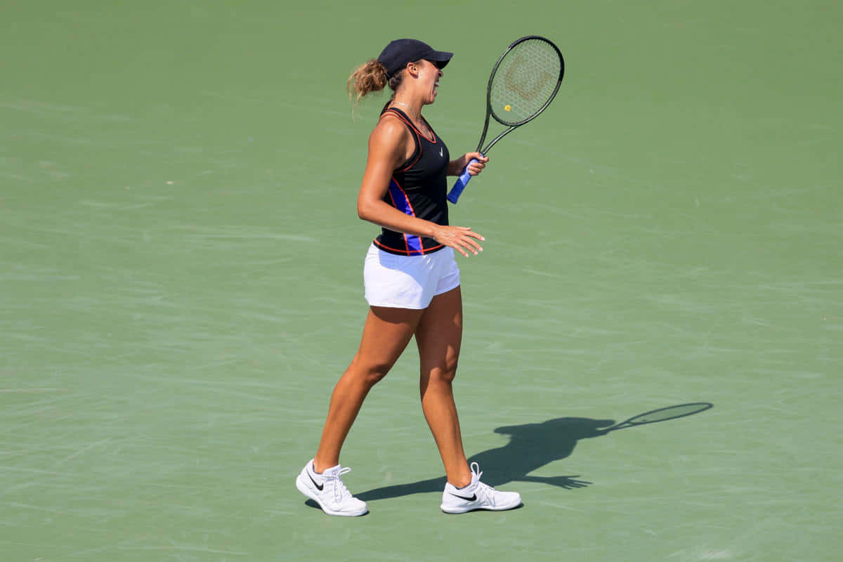 Athletic 桌布 壁紙 — 一位身穿黑色上衣和白色短褲的女網球運動員，自信地拿著球拍在綠色球場上大步走。