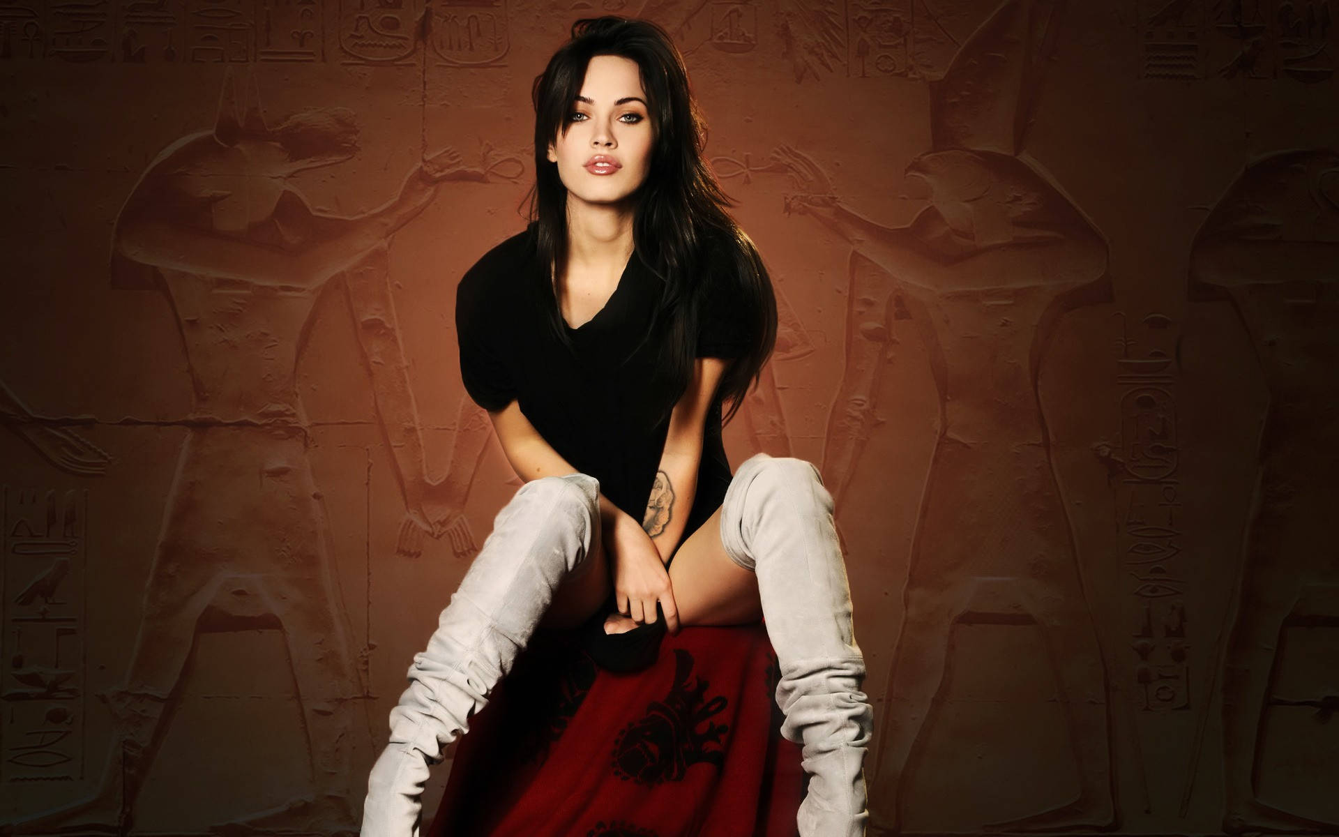 Megan Fox HD 桌布 壁紙 — 一位身穿黑色上衣和白色靴子的女性，自信地坐在紅色布幔王座上，背景是古埃及牆壁雕刻。