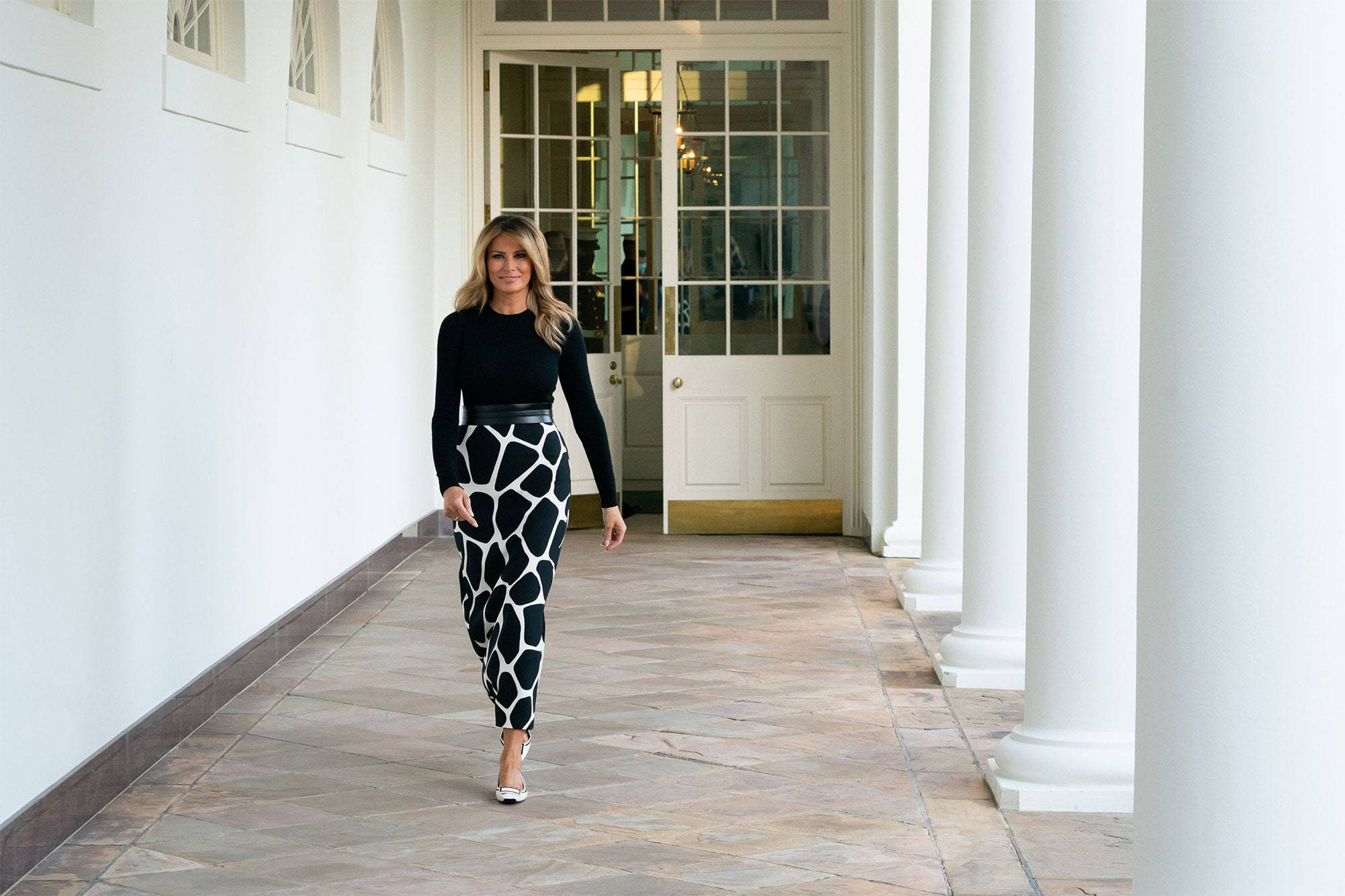 Melania 桌布 壁紙 — 一位身穿黑色上衣和長頸鹿紋褲子的女性自信地走在白色柱廊和瓷磚地板的走廊上。