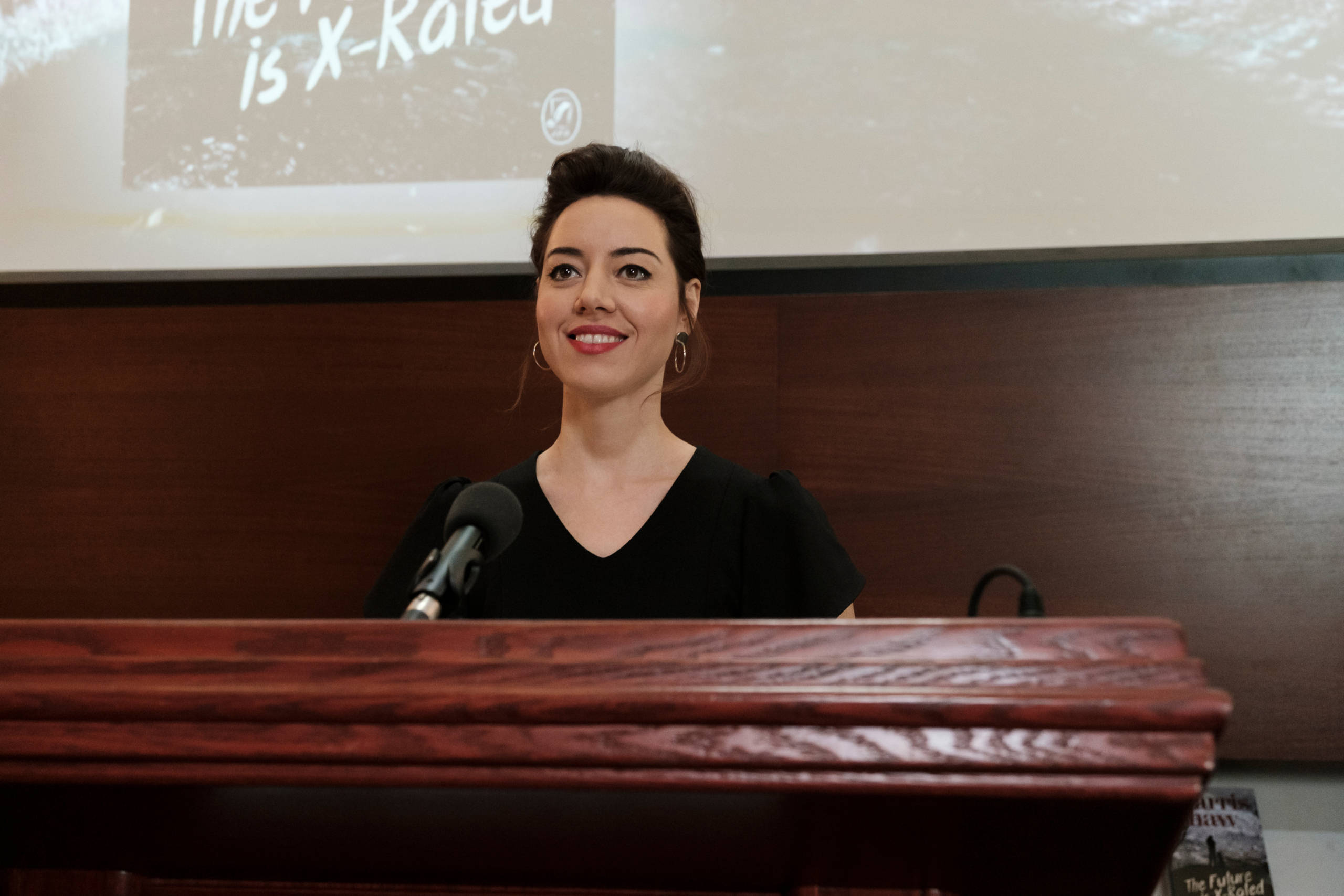 Aubrey 桌布 壁紙 — 一位身穿黑色上衣的女性站在拋光木製講台後面，手持麥克風，溫暖地微笑。