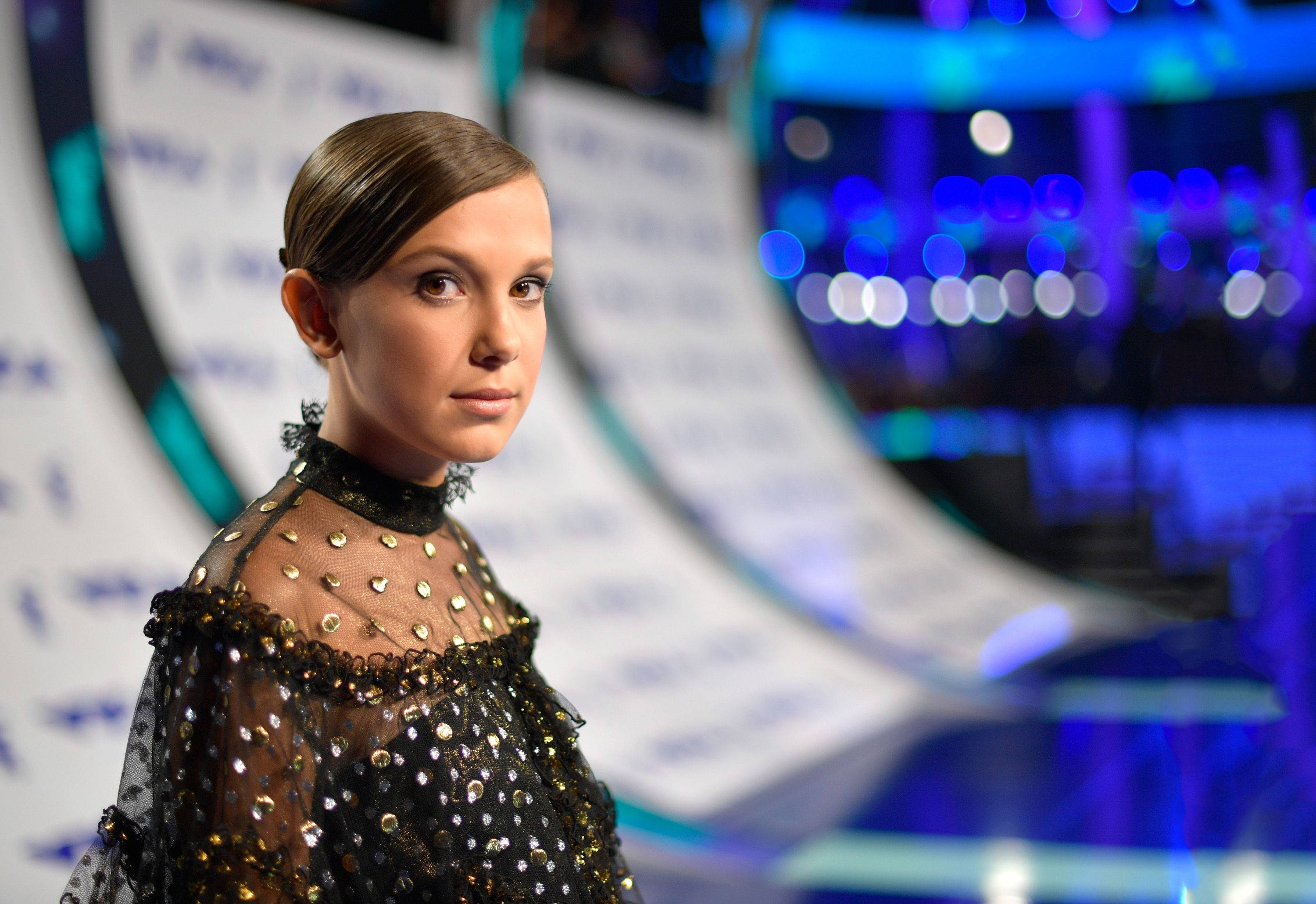 Millie 桌布 壁紙 — 一位身穿黑色亮片禮服、肩部透視的女性，在充滿活力的藍色散景舞台背景下向前凝視。