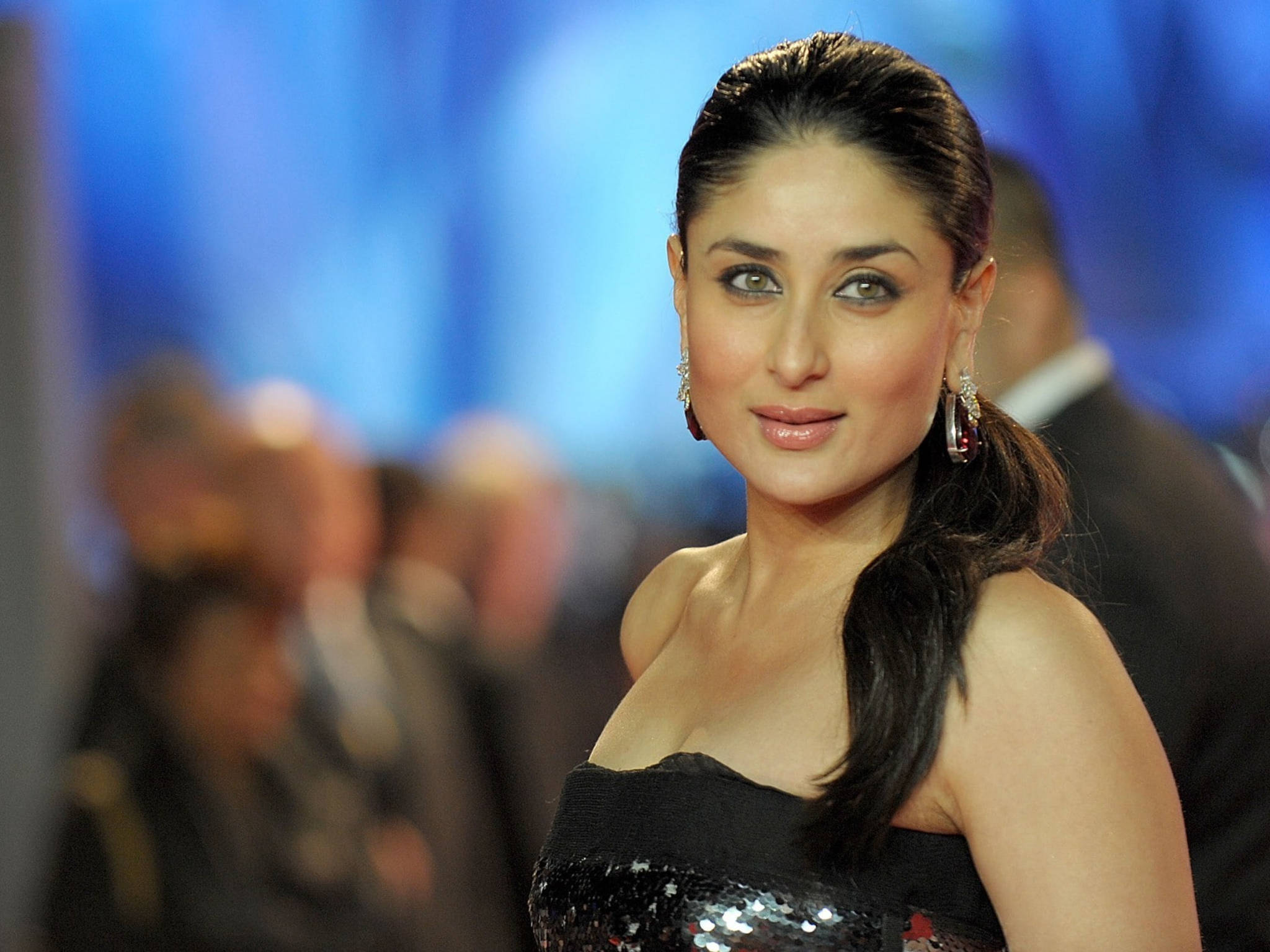 Kareena 桌布 壁紙 — 一位身穿黑色亮片連衣裙的女性在模糊的藍色活動背景前對著鏡頭微笑。