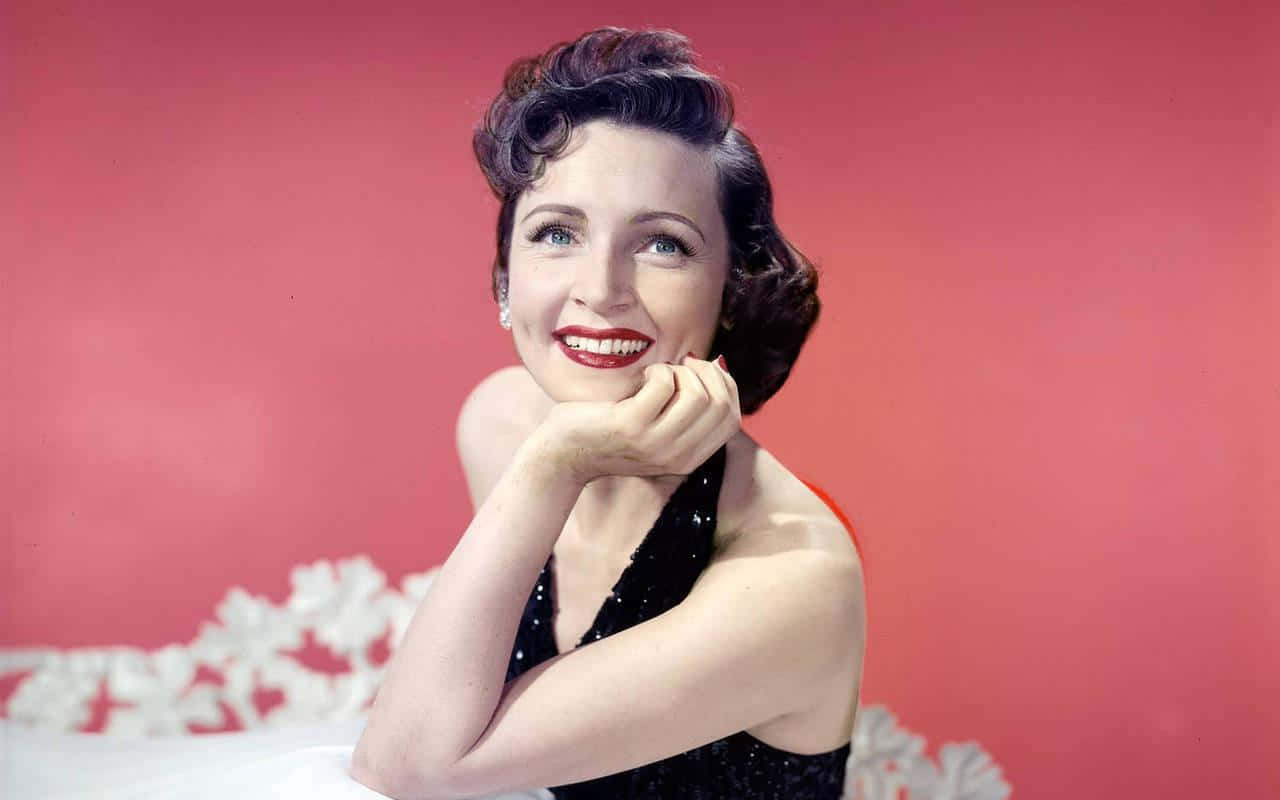 Betty White 桌布 壁紙 — 一位身穿黑色亮片連衣裙的迷人女性，在帶有白色花卉點綴的鮮豔粉紅色背景下燦爛微笑。