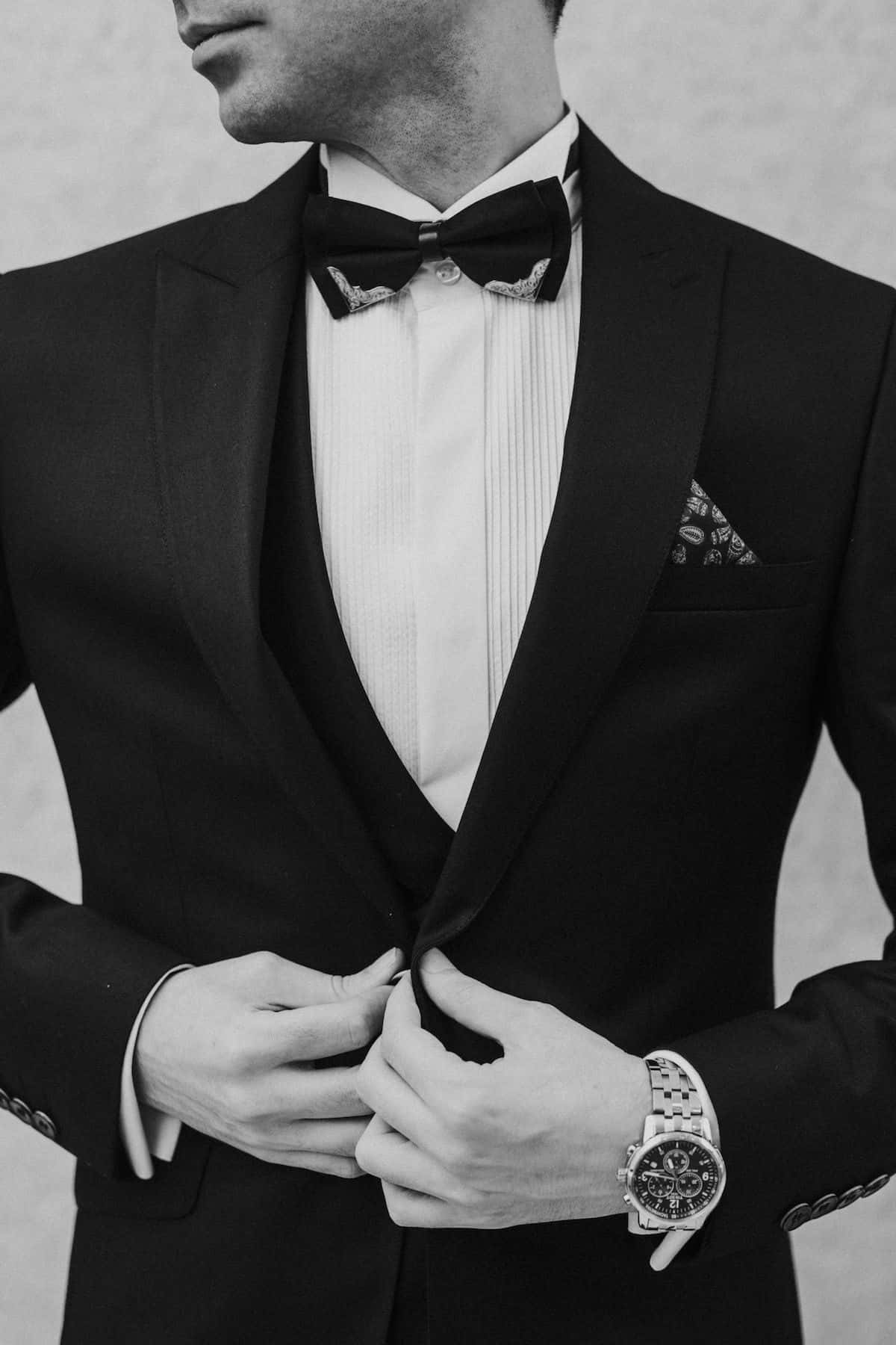 suit 桌布 壁紙 — 一位身穿黑色修身燕尾服的男士正在調整他的外套鈕扣，搭配圖案領結和口袋方巾。