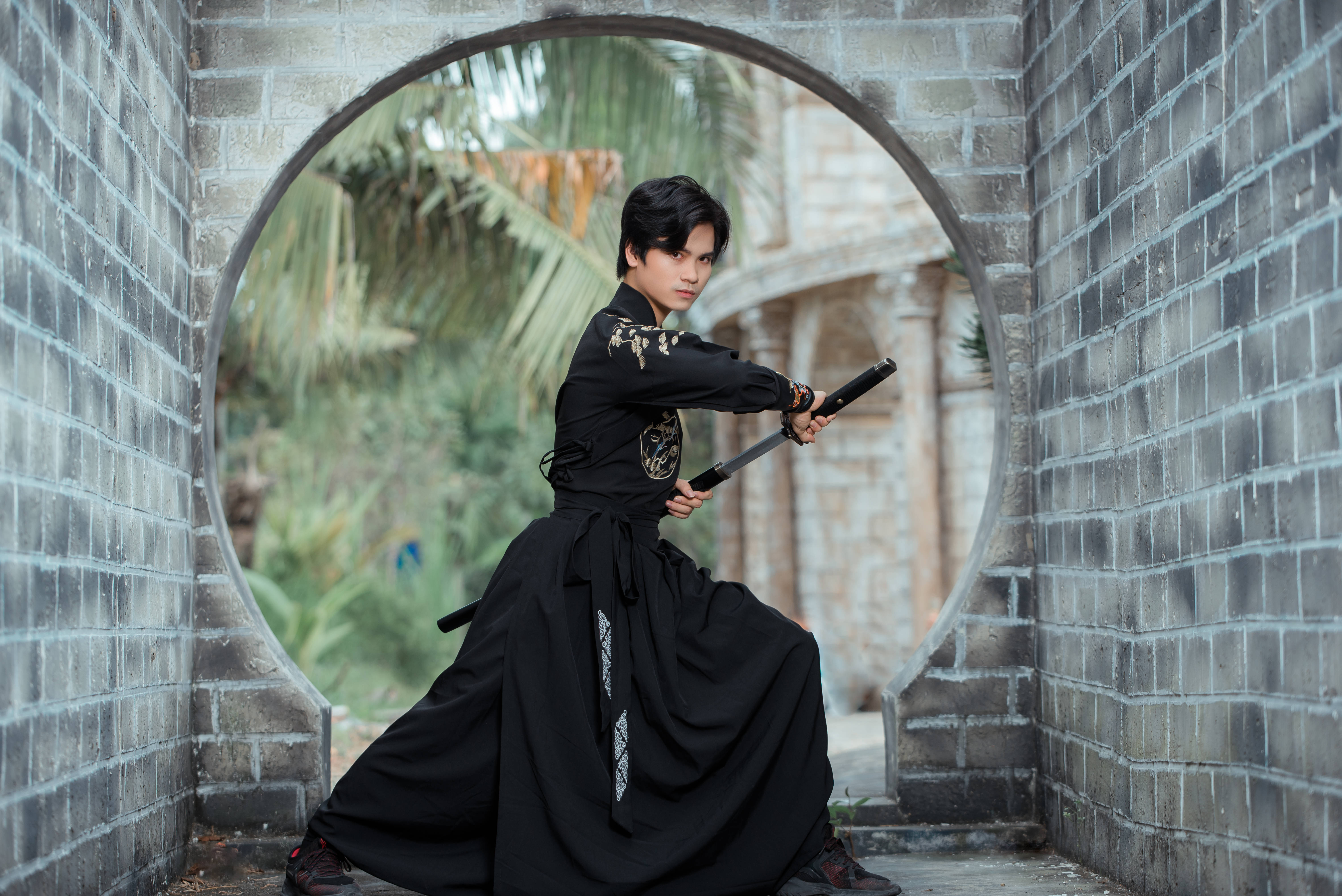 武士動畫 桌布 壁紙 — 一位身穿黑色傳統服飾的年輕戰士，在石拱門框下持劍而立，背景是茂密的綠植。