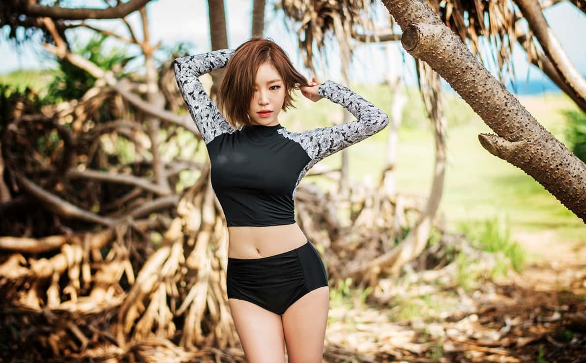 Brown Eyed Girl 桌布 壁紙 — 一位身穿黑色和圖案運動服的女性在陽光明媚的自然環境中，在纏結的樹根下自信地擺姿勢。