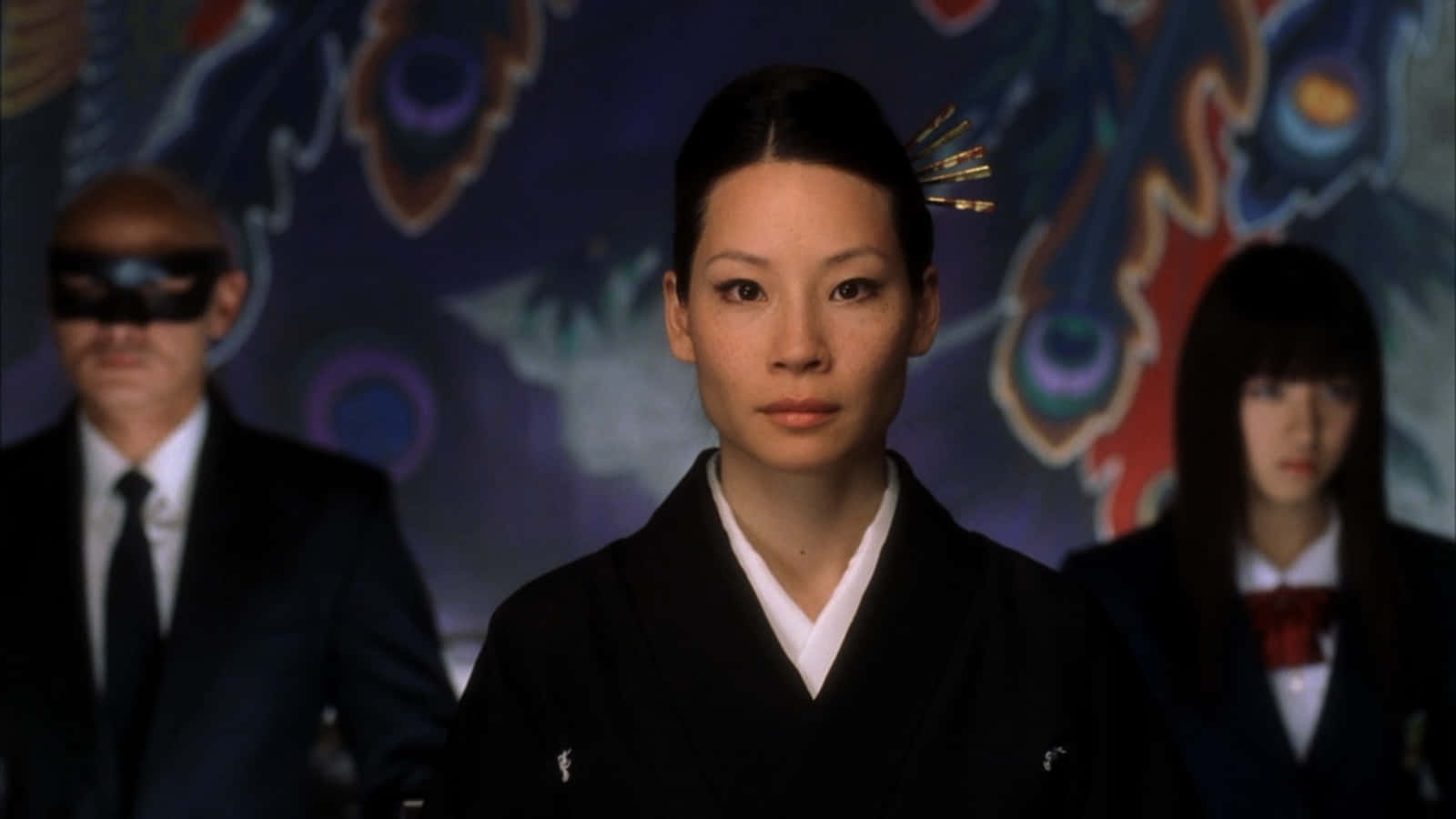 lucy liu 桌布 壁紙 — 一位身穿黑色和服的女性居中站立，身後有一位被蒙眼的男子和一位穿校服的女孩，背景是色彩繽紛的抽象圖案。
