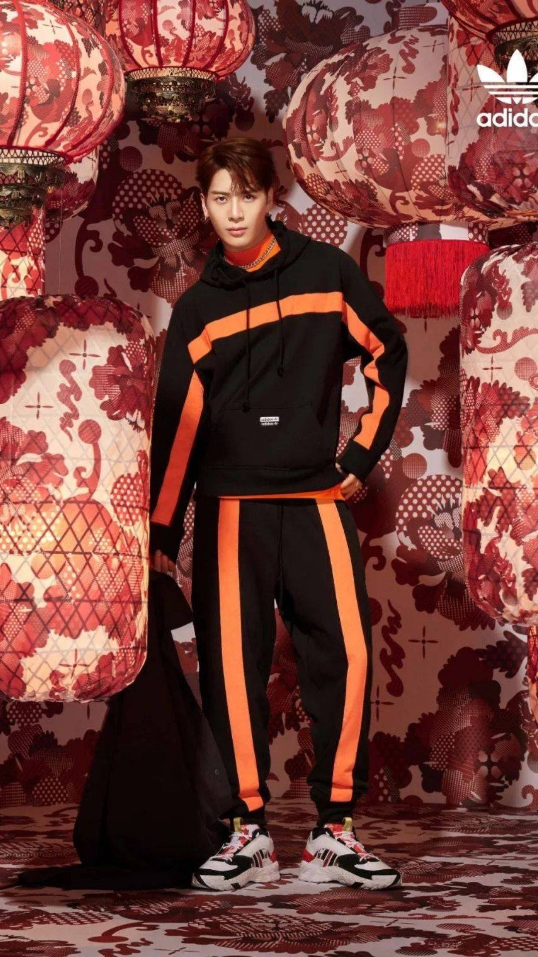 王嘉爾 桌布 壁紙 — 一位身穿黑色和橙色Adidas運動服的年輕男子自信地站在裝飾華麗的紅色燈籠之間，背景帶有圖案。
