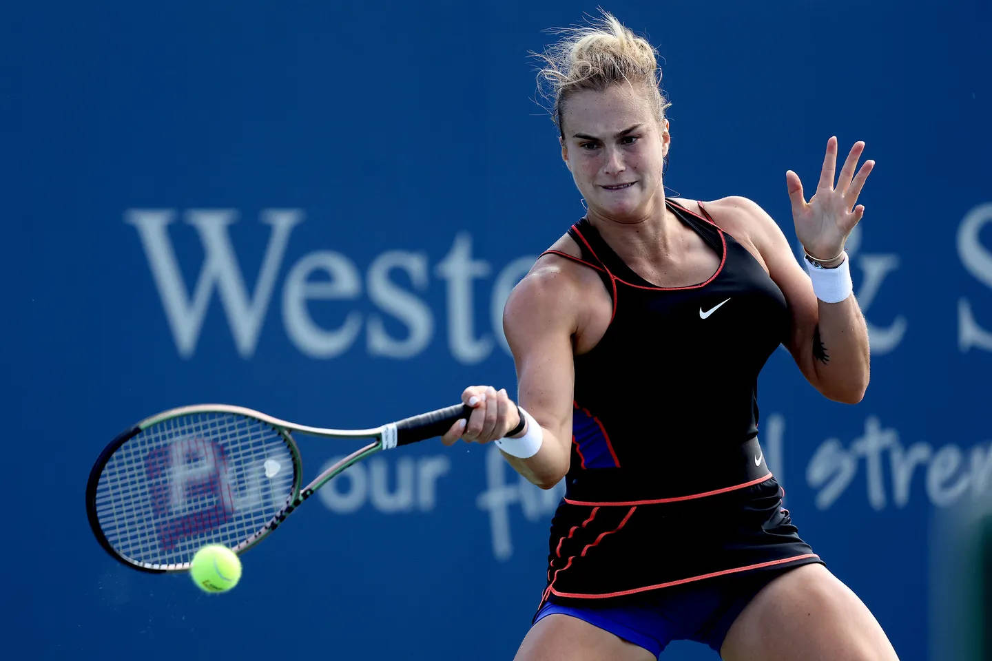 Aryna Sabalenka 桌布 壁紙 — 一位身穿黑色和藍色裝備的女網球運動員，在帶有白色文字的純藍色牆壁前揮拍擊打一個亮黃色的球。