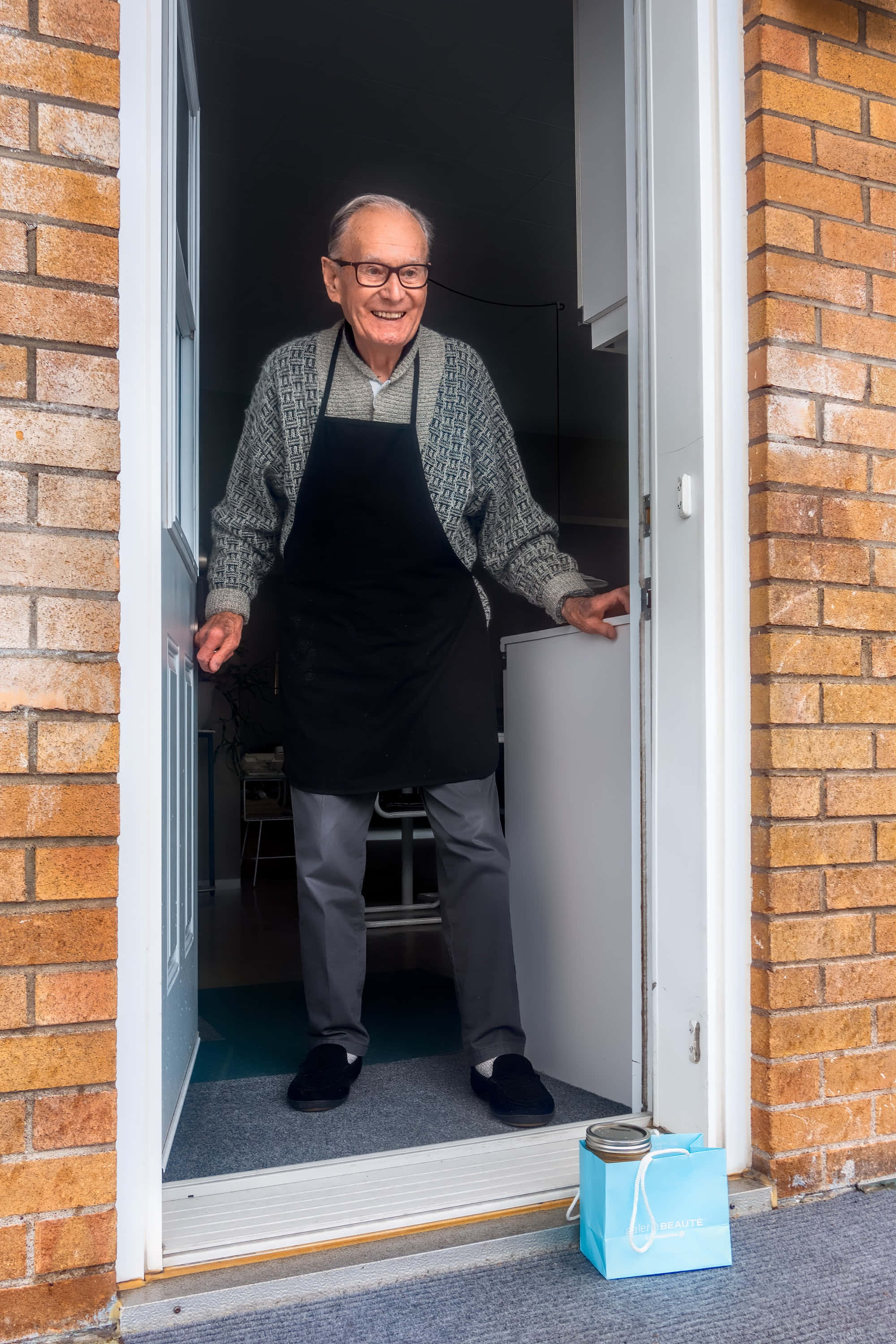 Mature 桌布 壁紙 — 一位身穿黑色圍裙的長者，微笑著穿過磚牆框著的門口。