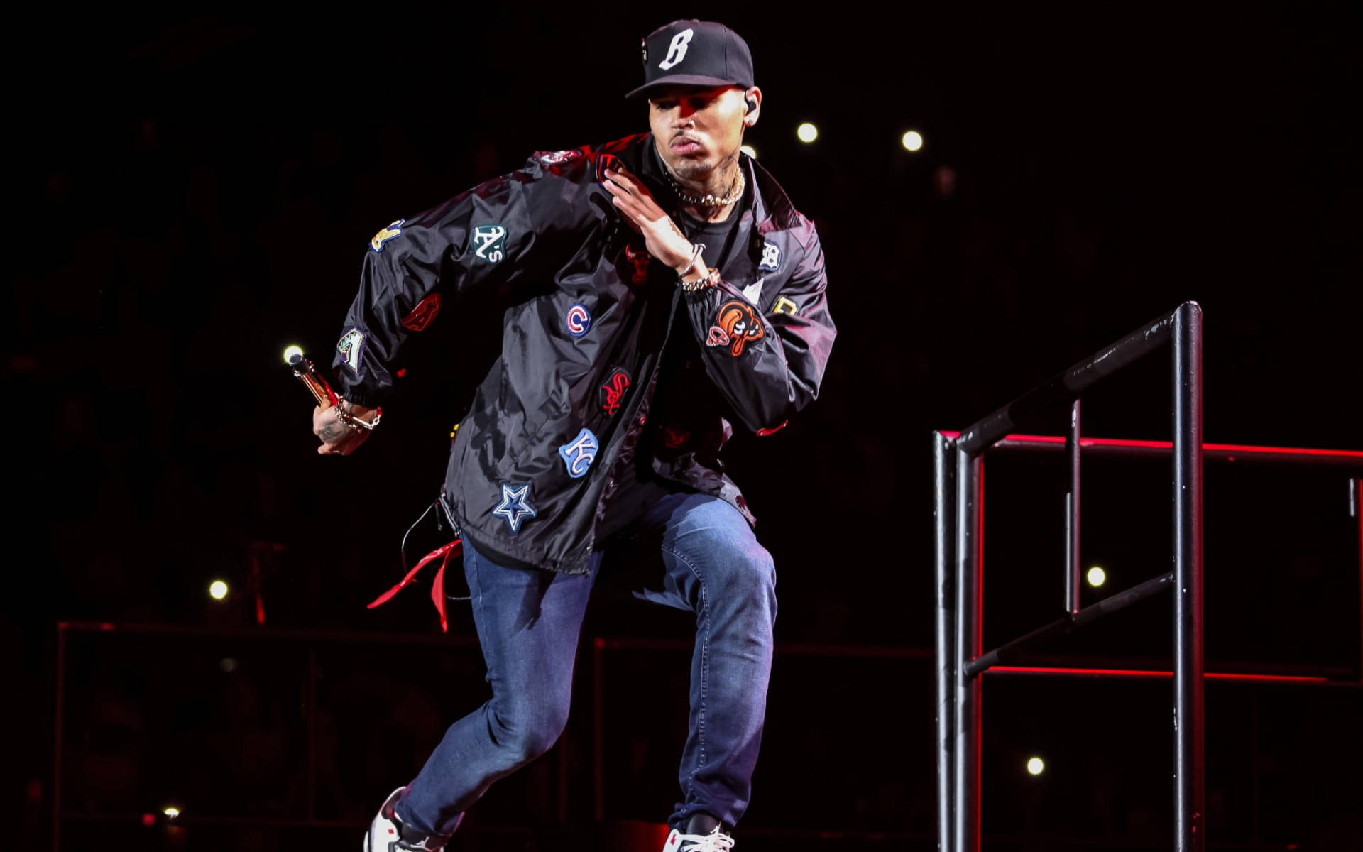 Chris Brown 桌布 壁紙 — 一位身穿黑色外套和帽子的男表演者在燈光昏暗、帶有紅色欄杆和聚光燈的舞台上動態跳躍。