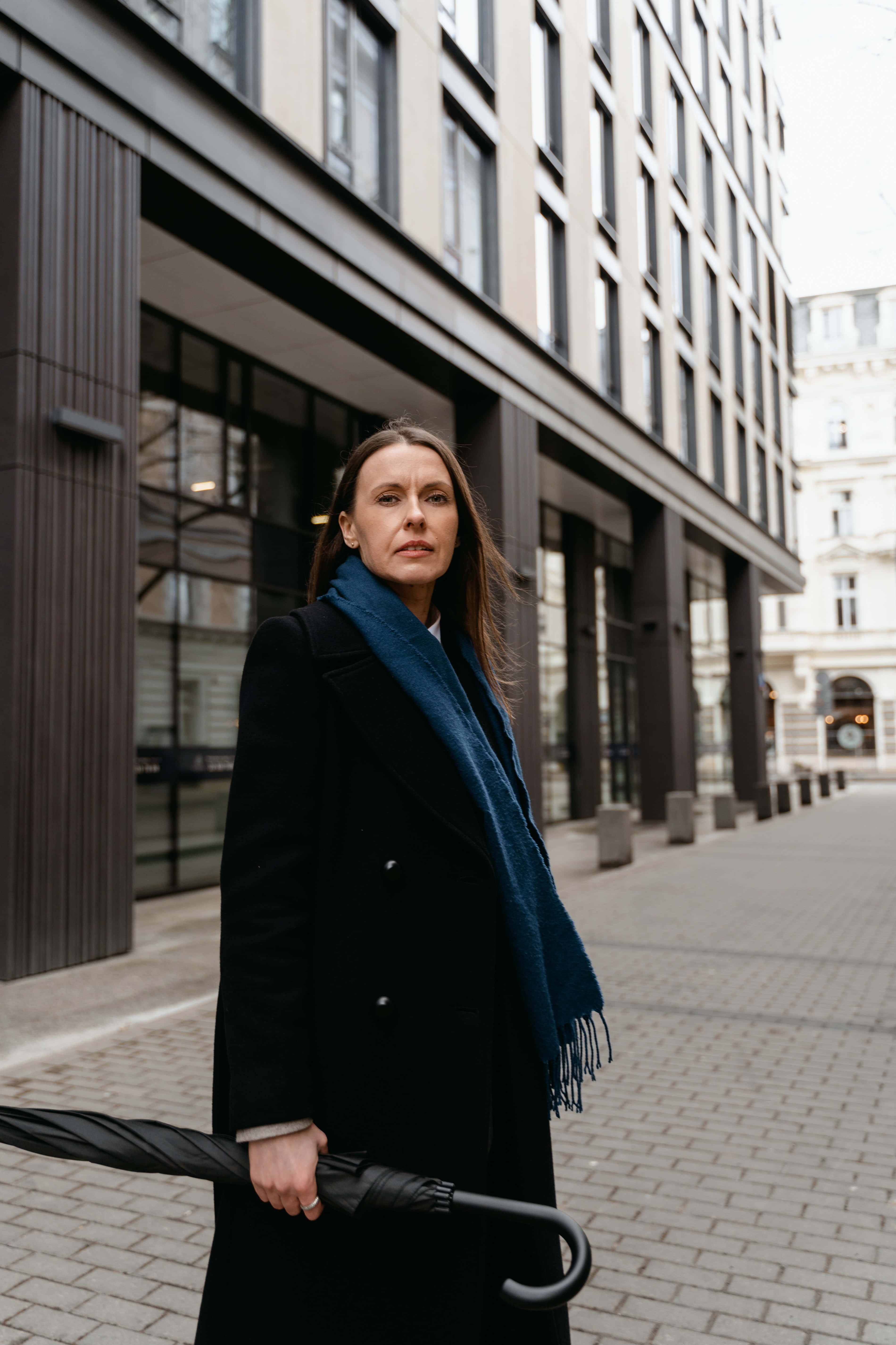 Navy Blue Scarf 桌布 壁紙 — 一位身穿黑色外套和藍色圍巾的女性站在城市人行道上，在陰天中拿著一把收起的雨傘。