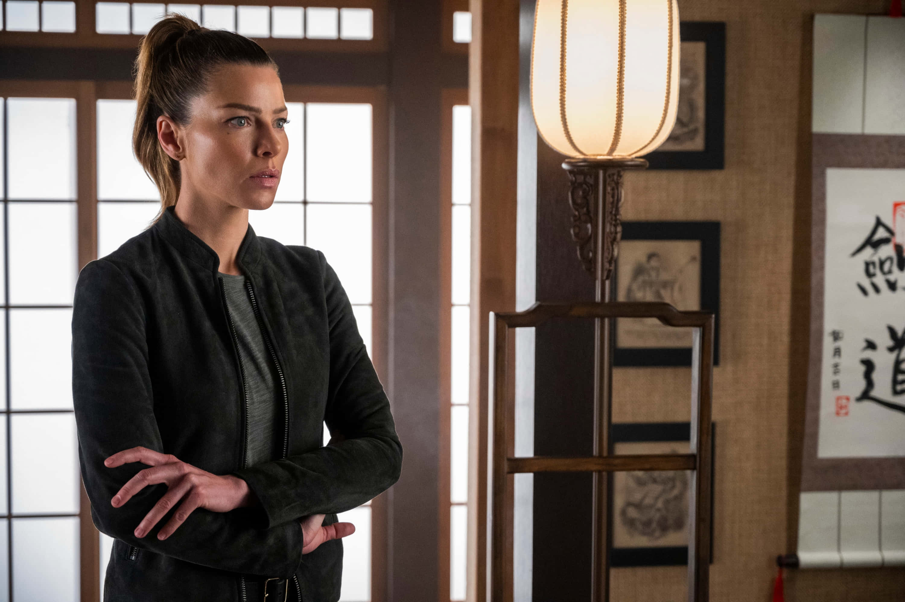 Lauren German 桌布 壁紙 — 一位身穿黑色外套的女性，在設有障子屏風和書法卷軸的傳統日式房間中交叉雙臂站立。