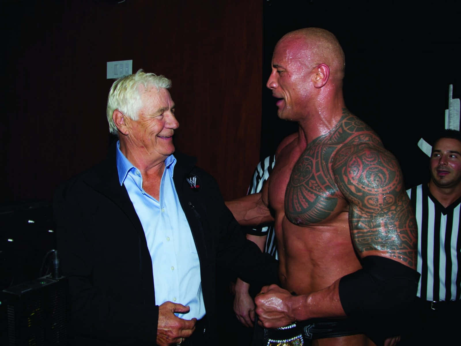 The Rock 桌布 壁紙 — 一位身穿黑色外套的年長男性在燈光昏暗的後台區域，對一位肌肉發達、滿身刺青的摔角手露出溫暖的笑容。