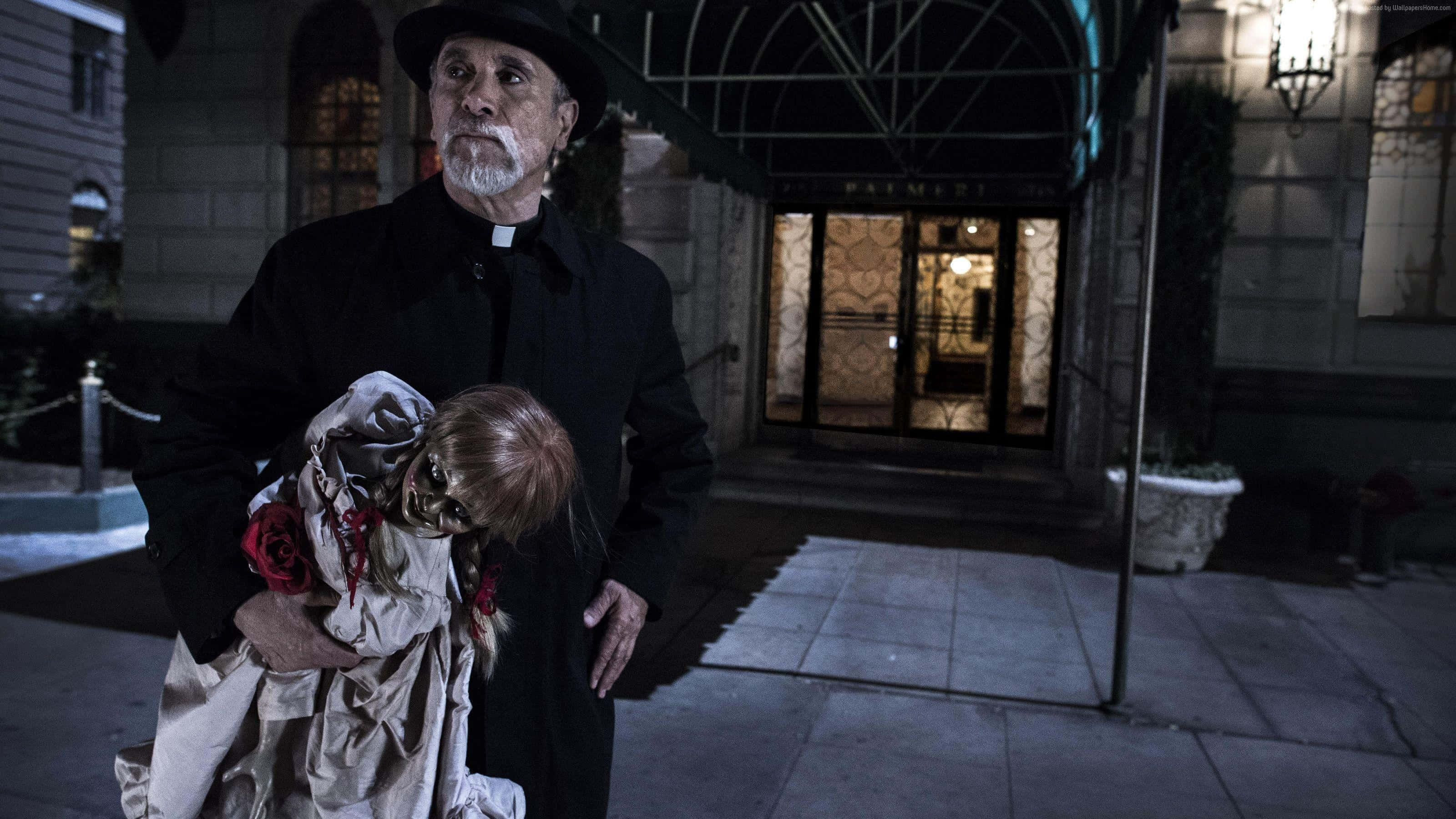 Annabelle 桌布 壁紙 — 一位身穿黑色外套的牧師在夜晚手持一個帶有紅玫瑰的血腥玩偶，站在燈光明亮的建築入口外。