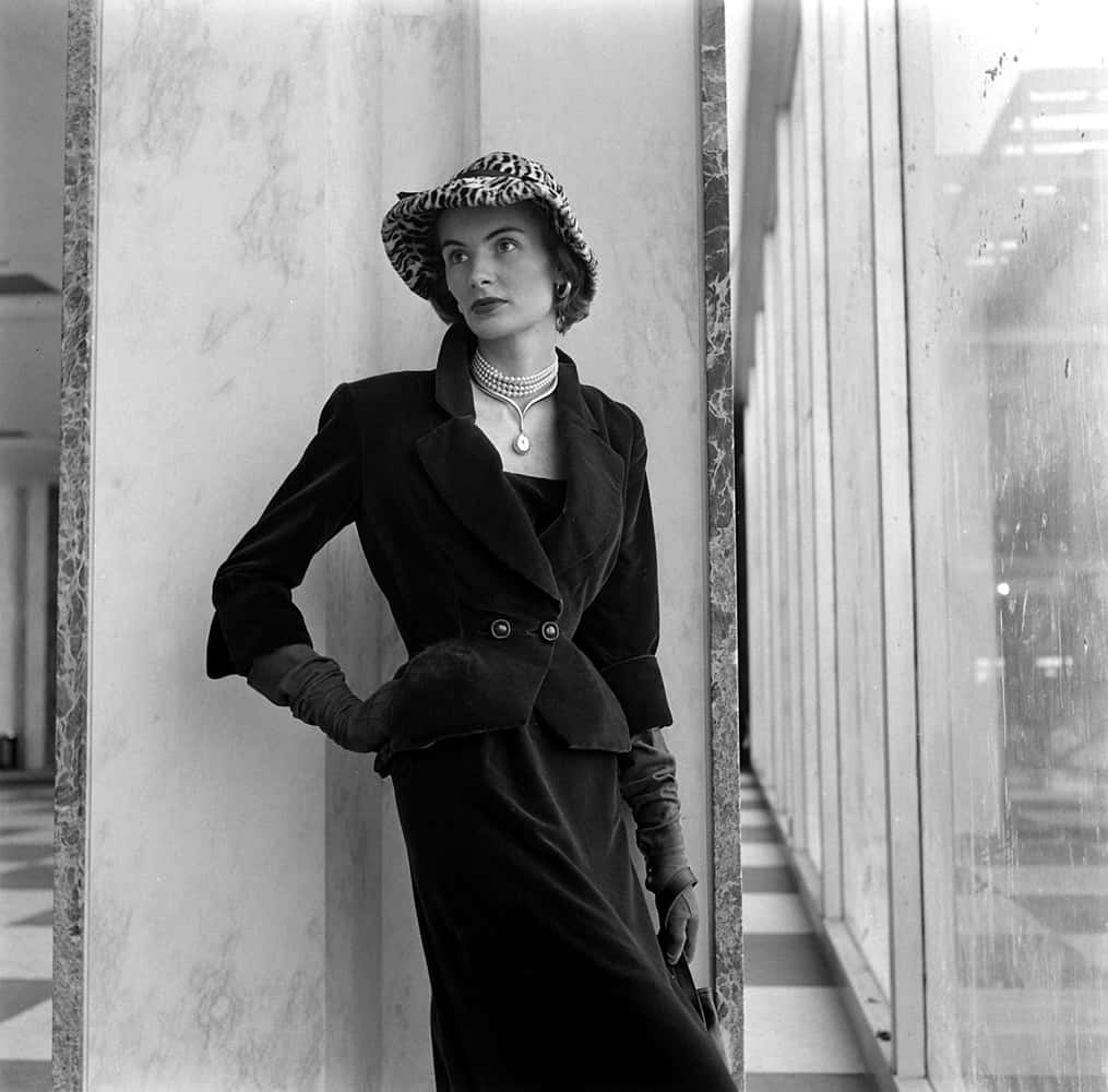 1950s 桌布 壁紙 — 一位身穿黑色天鵝絨西裝和豹紋帽子的優雅女士，倚靠在時尚現代走廊的大理石柱上。