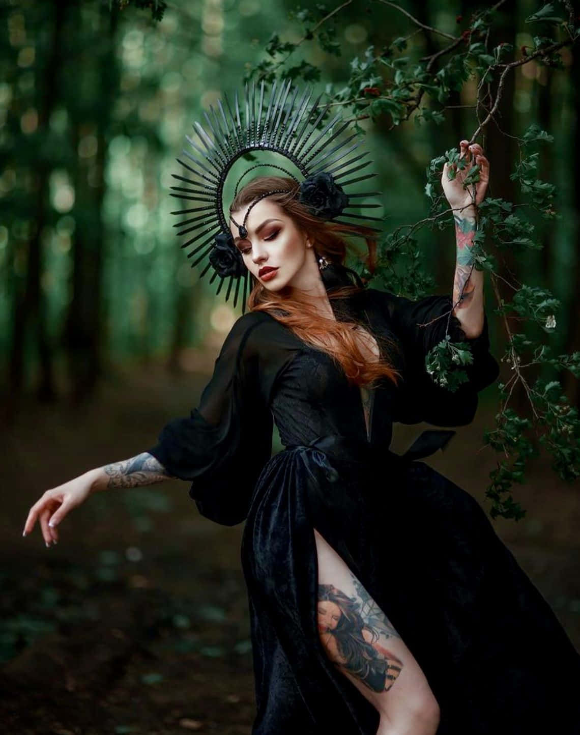 Witch Costumes 桌布 壁紙 — 一位身穿黑色天鵝絨長袍、頭戴尖刺光環皇冠的女子，優雅地站在鬱鬱蔥蔥的綠色森林中，她紋身的手臂伸向藤蔓。