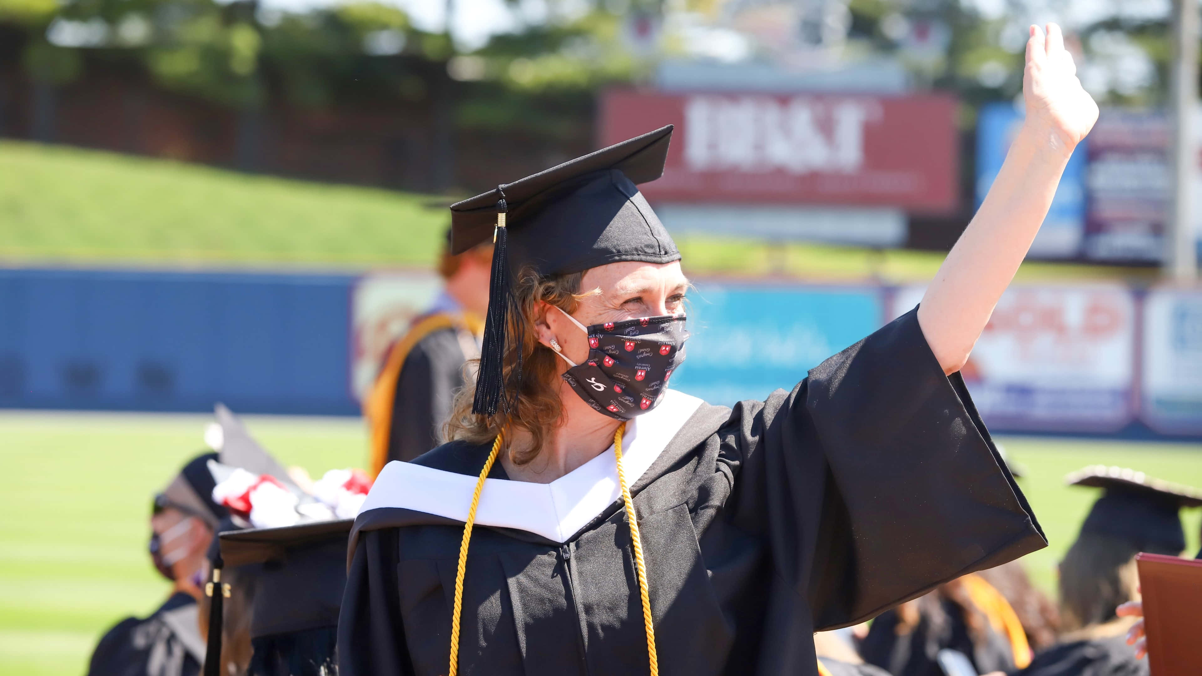 Graduation 桌布 壁紙 — 一位身穿黑色學士袍和畢業帽的畢業生，在戶外典禮上戴著圖案口罩，愉快地揮手。