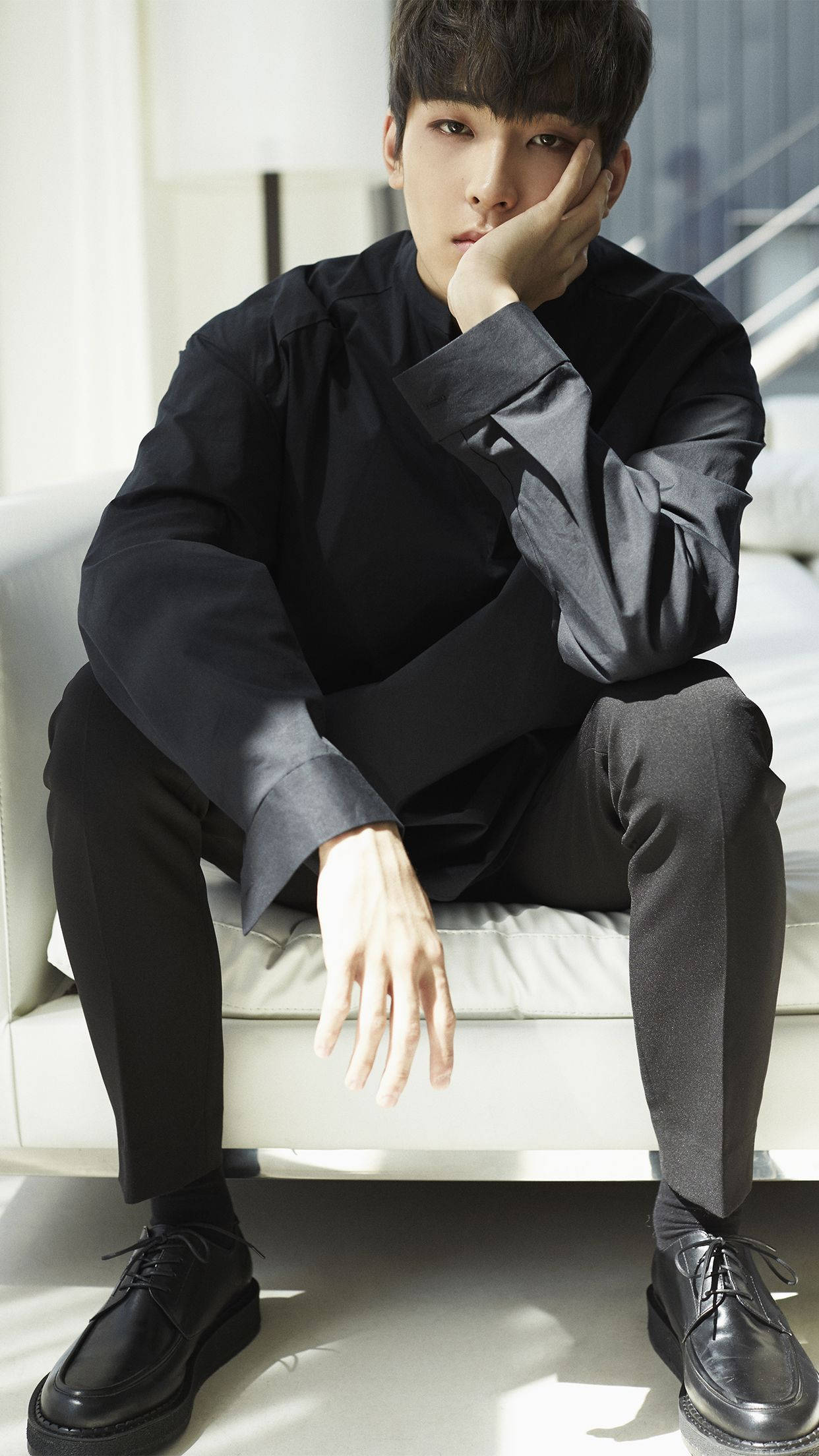 Wonwoo 桌布 壁紙 — 一位身穿黑色服裝的年輕男子沉思地坐在白色沙發上，陽光在地板上投下柔和陰影。