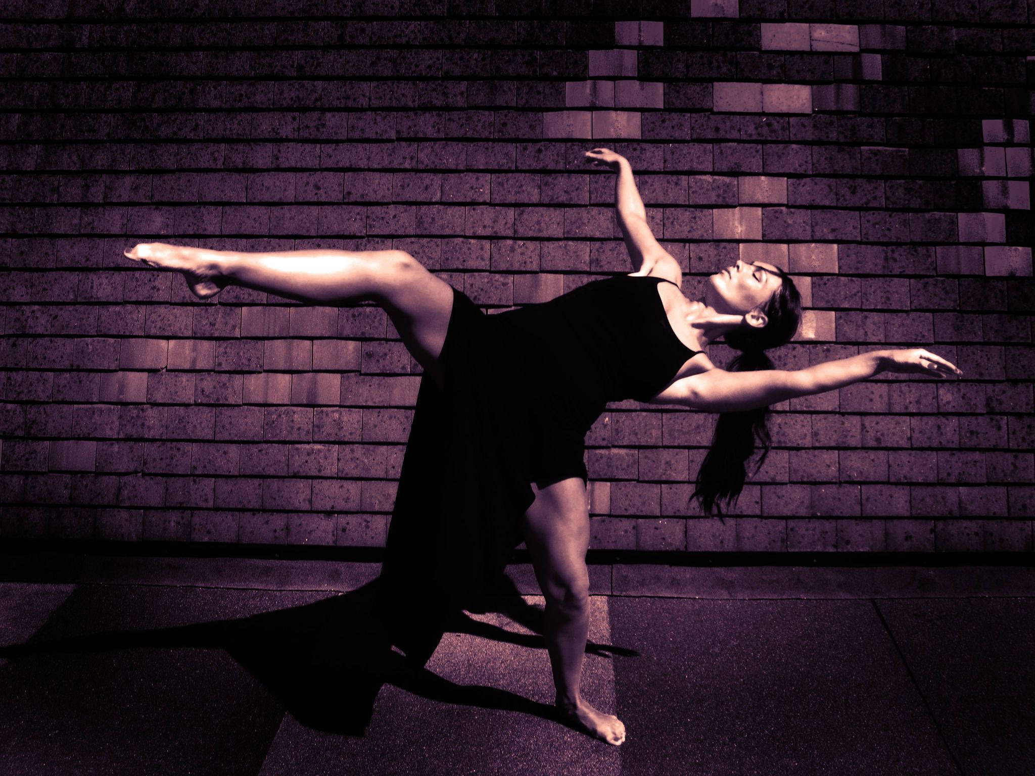 dancer 桌布 壁紙 — 一位身穿黑色服裝的舞者在紫色燈光的磚牆前擺出優雅的姿勢，投下長長的影子。