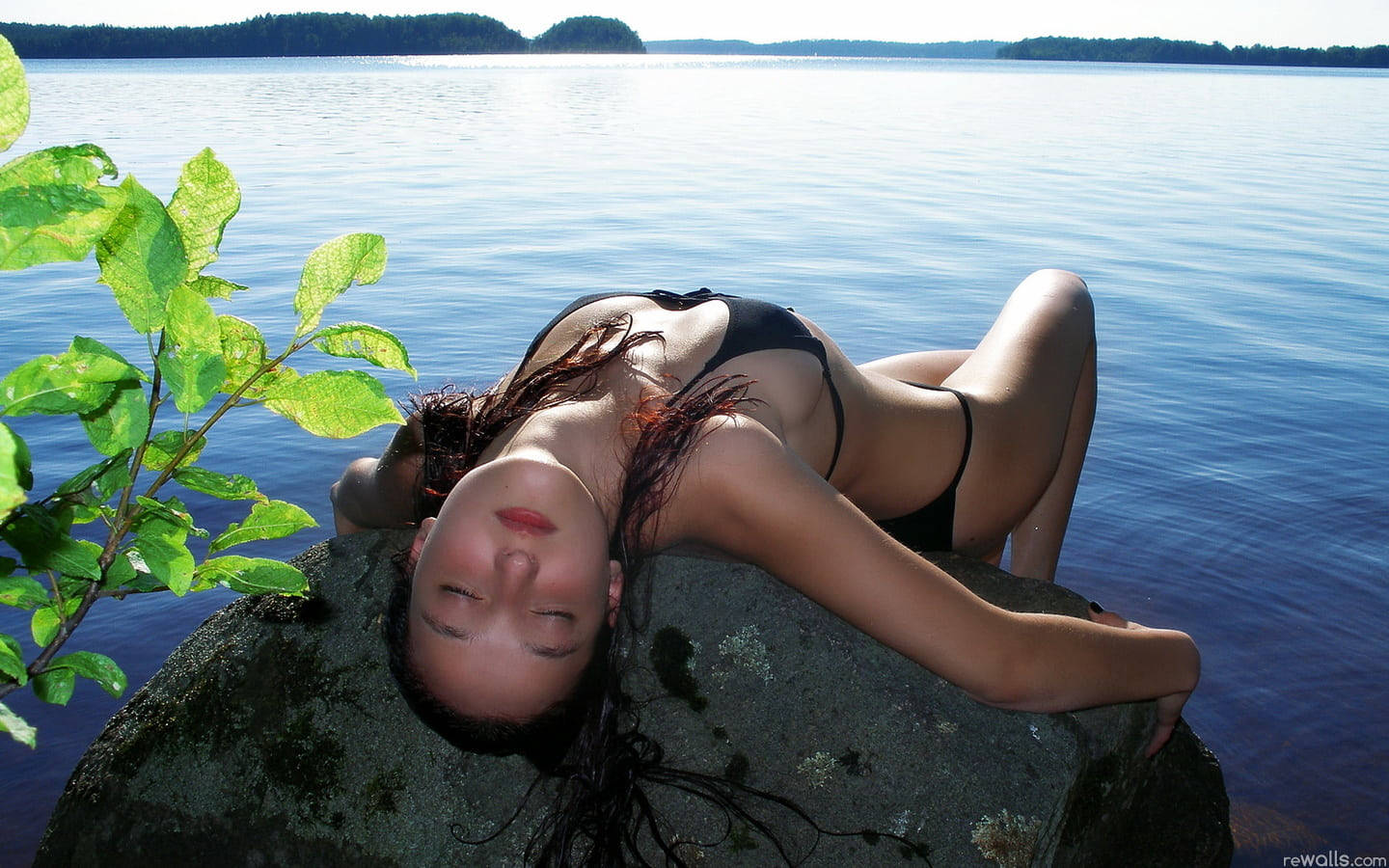 sexy bikini 桌布 壁紙 — 一位身穿黑色比基尼的女性在平靜的藍色湖邊，將頭靠在長滿苔蘚的石頭上，綠色樹葉構成了前景。