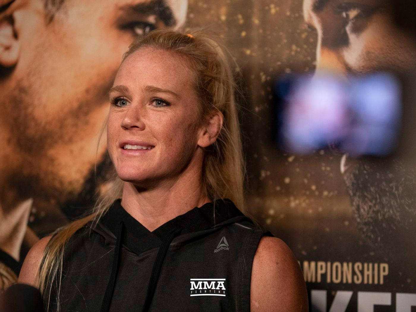 MMA 桌布 壁紙 — 一位身穿黑色無袖連帽衫的女性MMA拳手在記者會上溫柔微笑，身後有冠軍海報。
