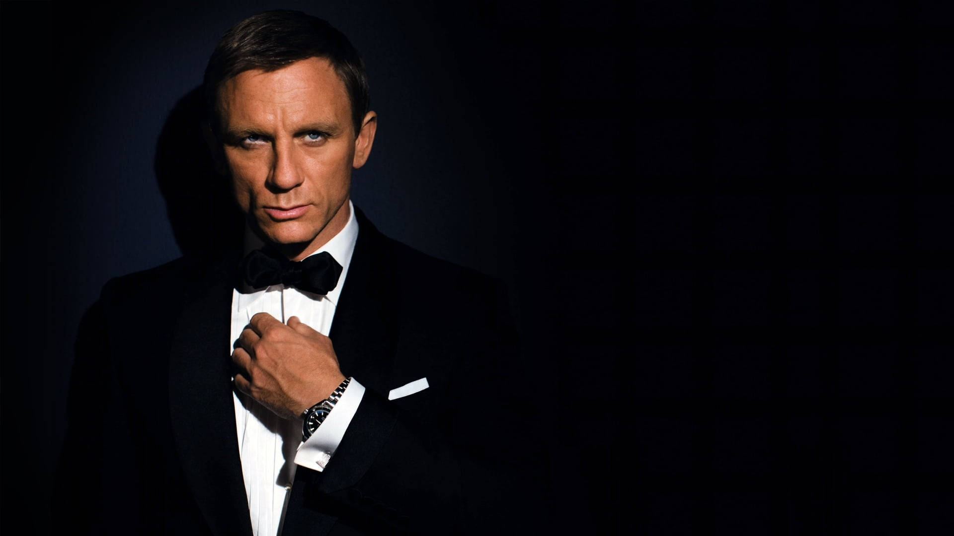 Daniel Craig 桌布 壁紙 — 一位身穿黑色燕尾服、打著蝴蝶結的男子，在黑暗背景下，手調整著西裝領，燈光銳利。