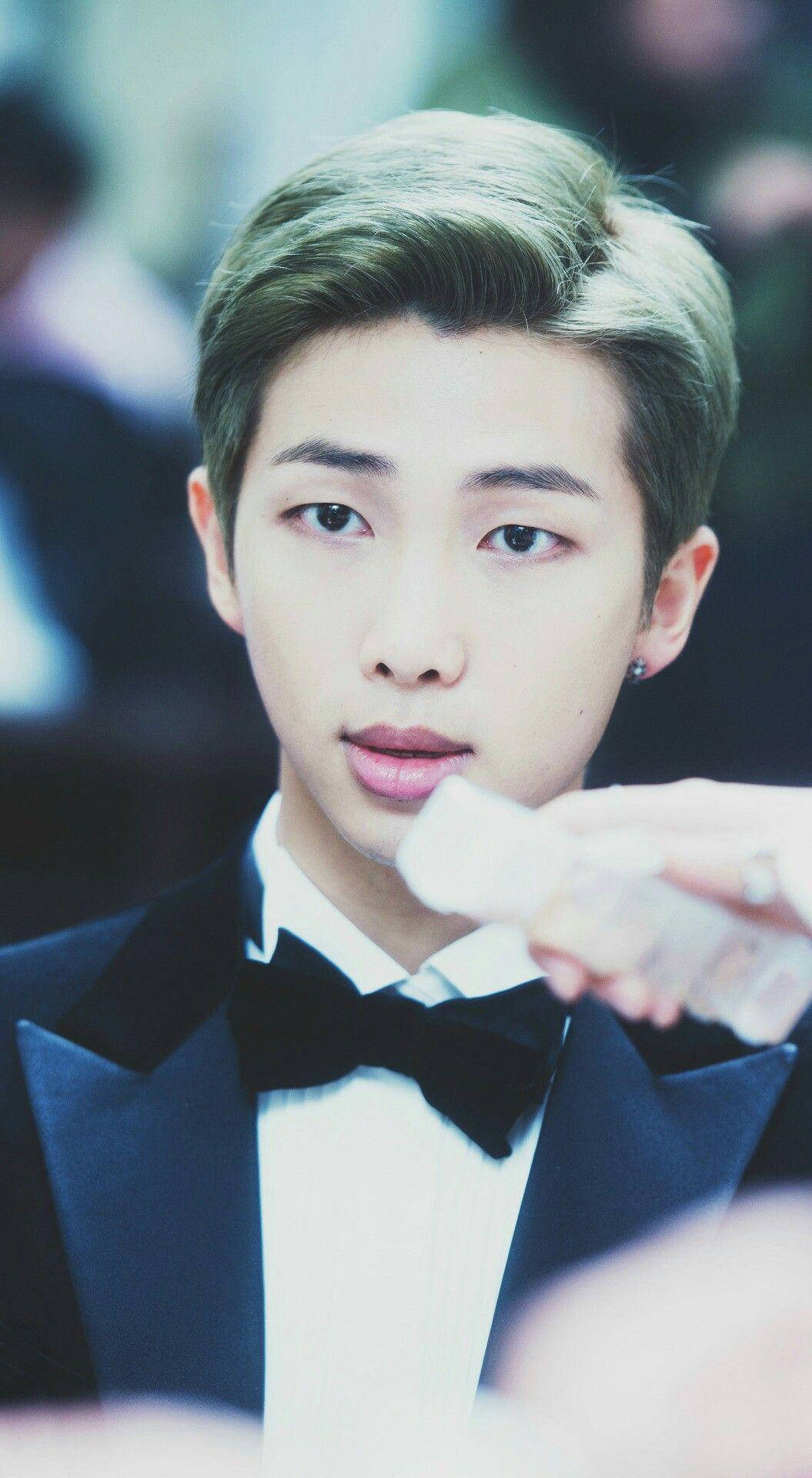 Kpop Aesthetic 桌布 壁紙 — 一位身穿黑色燕尾服、打著領結的年輕男子，手持透明塑膠杯，冷靜地直視鏡頭。