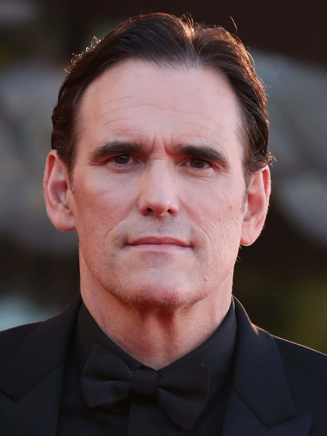 matt dillon 桌布 壁紙 — 一位身穿黑色燕尾服和領結、表情嚴肅的男士在溫暖燈光下的特寫肖像。