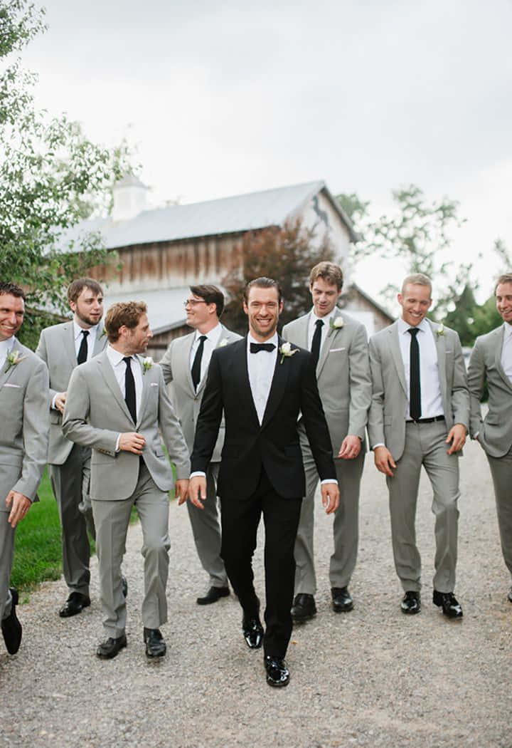 Groomsmen 桌布 壁紙 — 一位身穿黑色燕尾服的新郎與他穿著灰色西裝、面帶微笑的伴郎們，愉快地走在靠近鄉村穀倉的碎石路上。