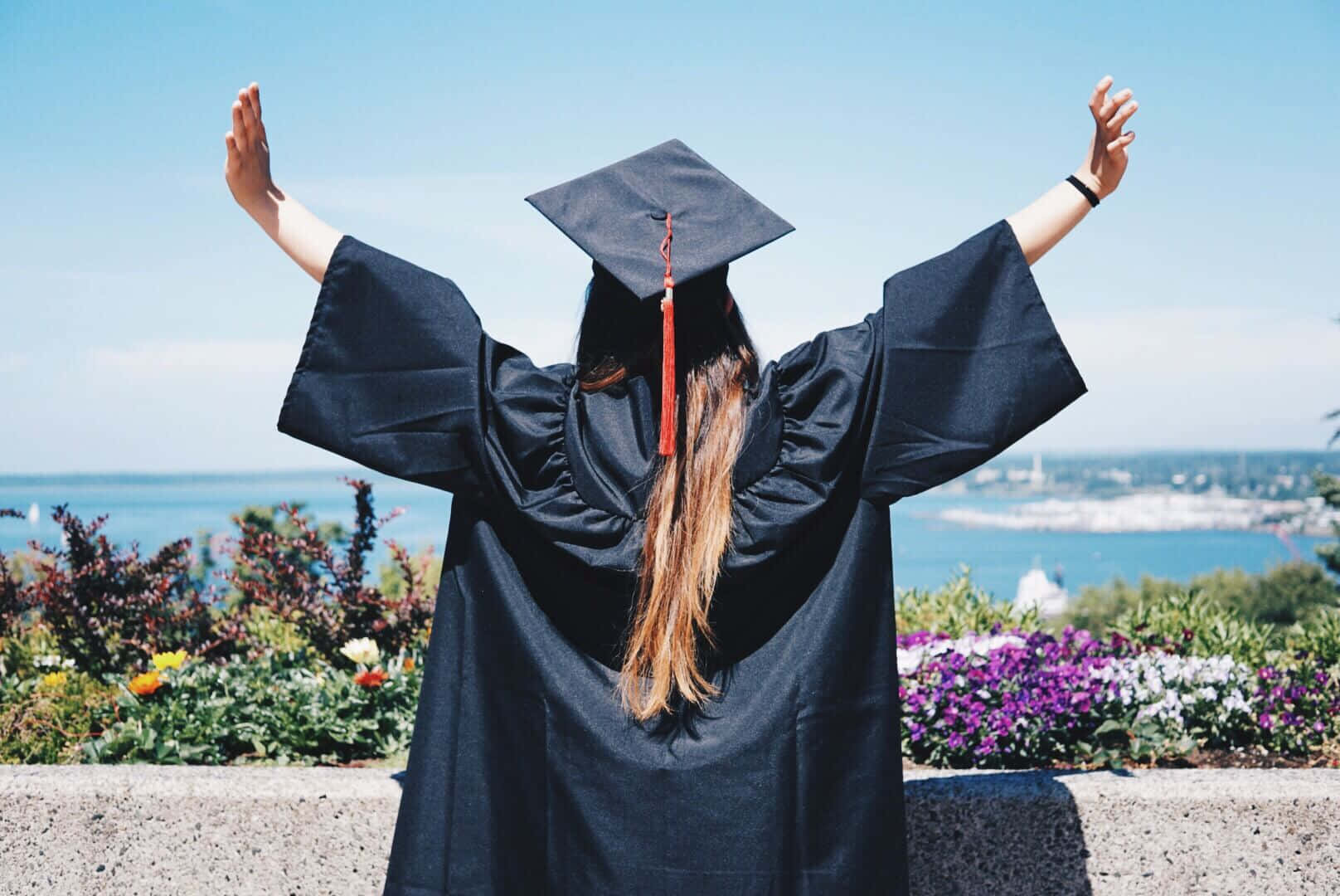 女孩 桌布 壁紙 — 一位身穿黑色畢業袍和四方帽的畢業生，高舉雙臂，以勝利姿態俯瞰著晴朗藍天下的沿海城市景觀。