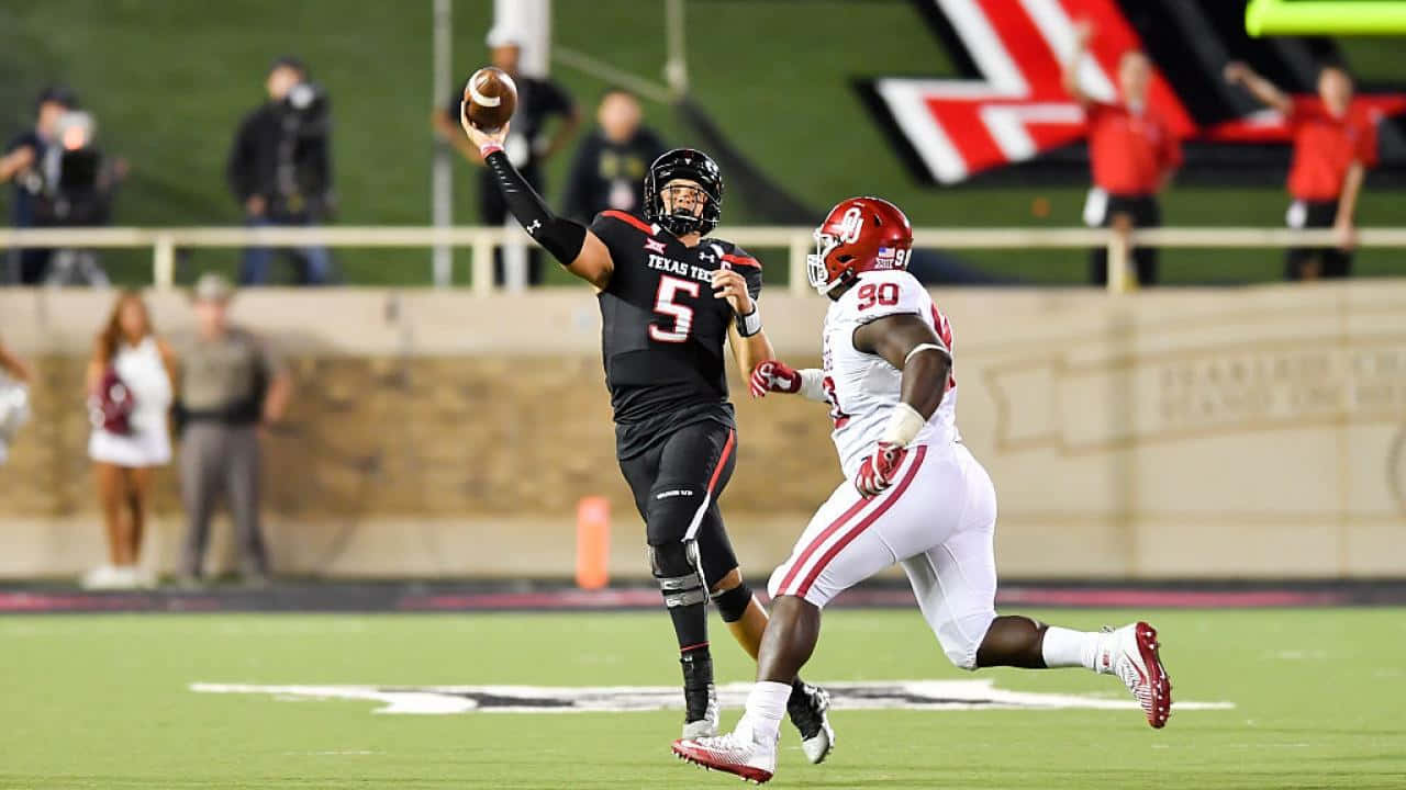 Texas Tech 桌布 壁紙 — 一位身穿黑色的德州理工四分衛擲出橄欖球，而一位戴紅色頭盔、穿白色的防守員在燈光球場上衝刺擒抱他。