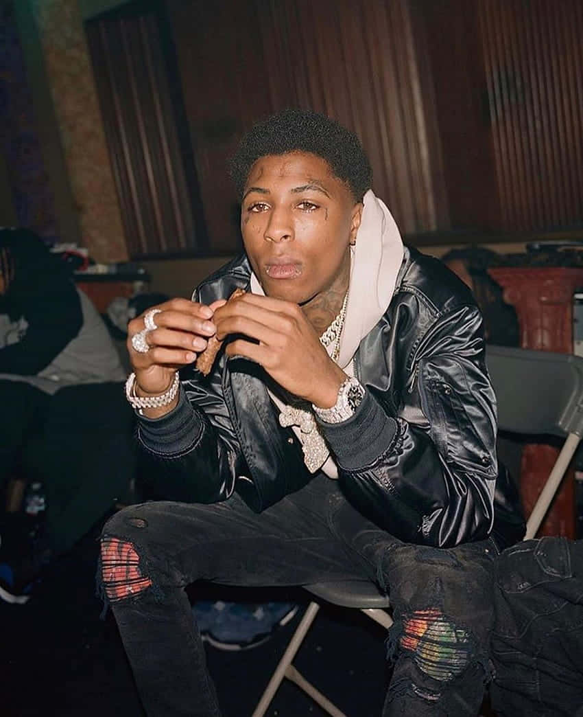 Youngboy 桌布 壁紙 — 一位身穿黑色皮夾克和破洞牛仔褲的年輕男子坐在折疊椅上，手持電話，表情嚴肅。