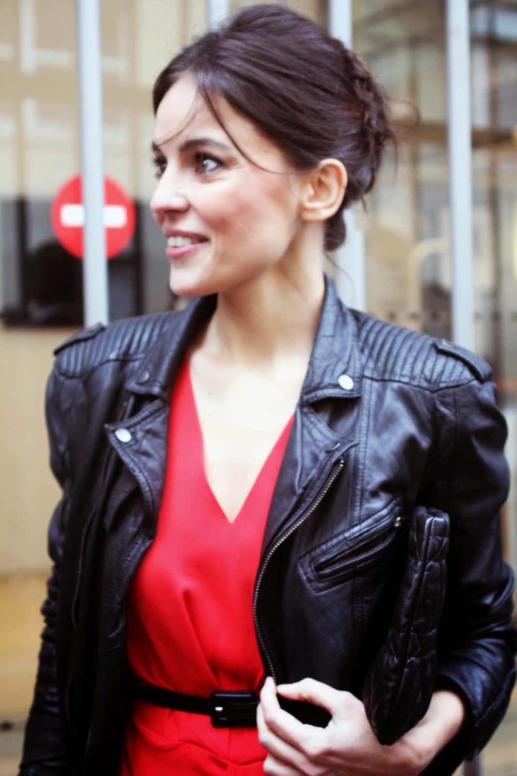 Elena Anaya 桌布 壁紙 — 一位身穿黑色皮夾克和紅色連衣裙的女性，在玻璃建築附近戶外手持手拿包微笑。