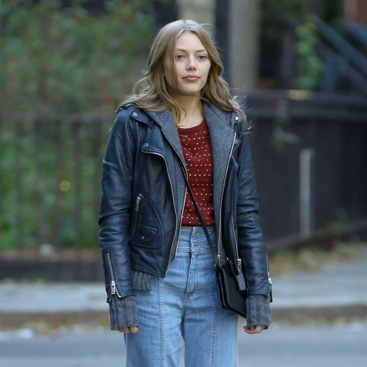 Grace Van Dien 桌布 壁紙 — 一位身穿黑色皮夾克和藍色牛仔褲的年輕女性自信地走在城市街道上，周圍散落著秋天的落葉。