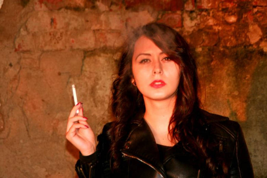 Smoking 桌布 壁紙 — 一位身穿黑色皮夾克的女性在溫暖燈光下，手持香煙靠著紋理磚牆。
