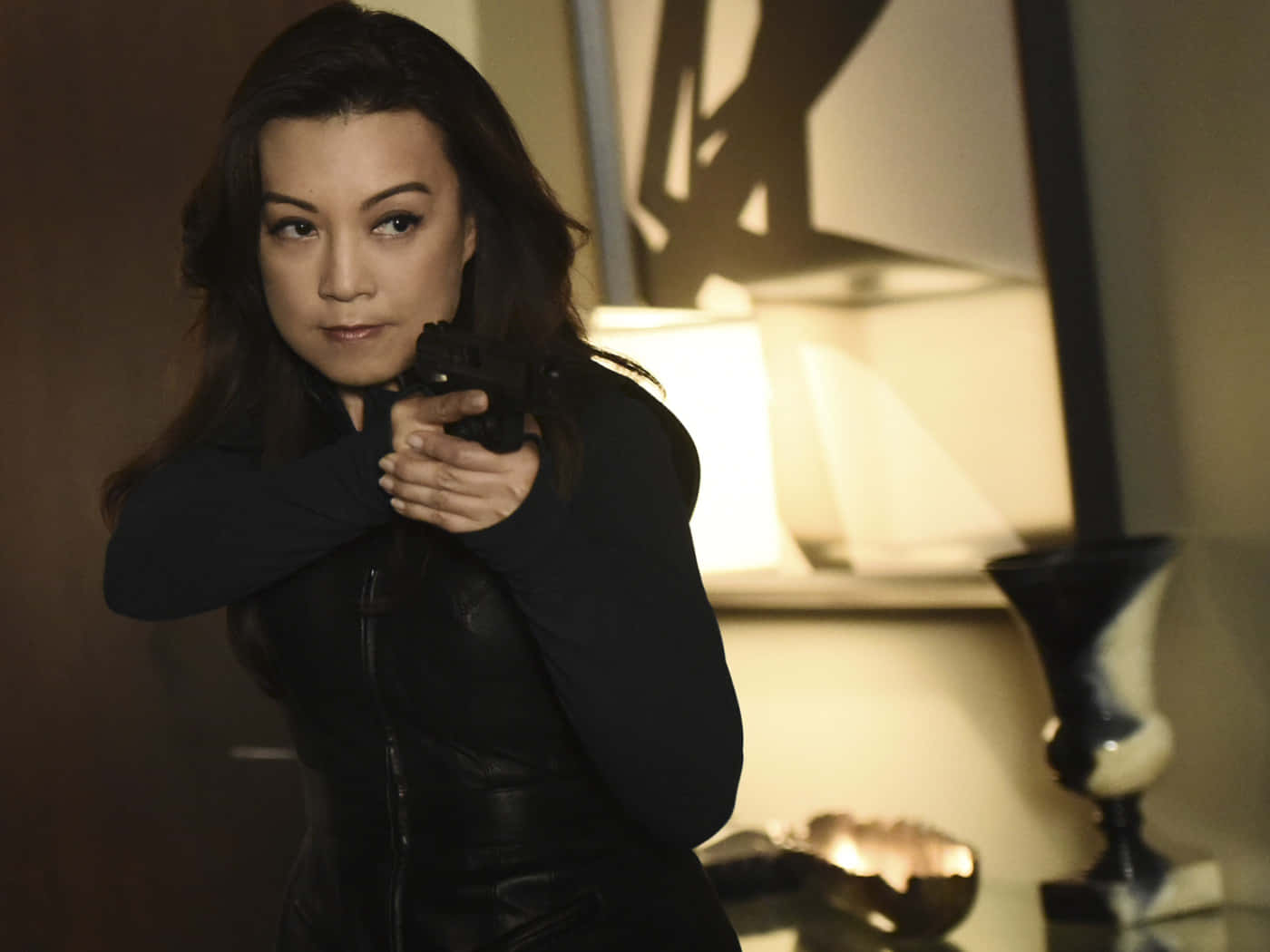 Ming Na Wen 桌布 壁紙 — 一位身穿黑色皮夾克的女性在溫暖燈光的室內環境中，專注地瞄準手槍。