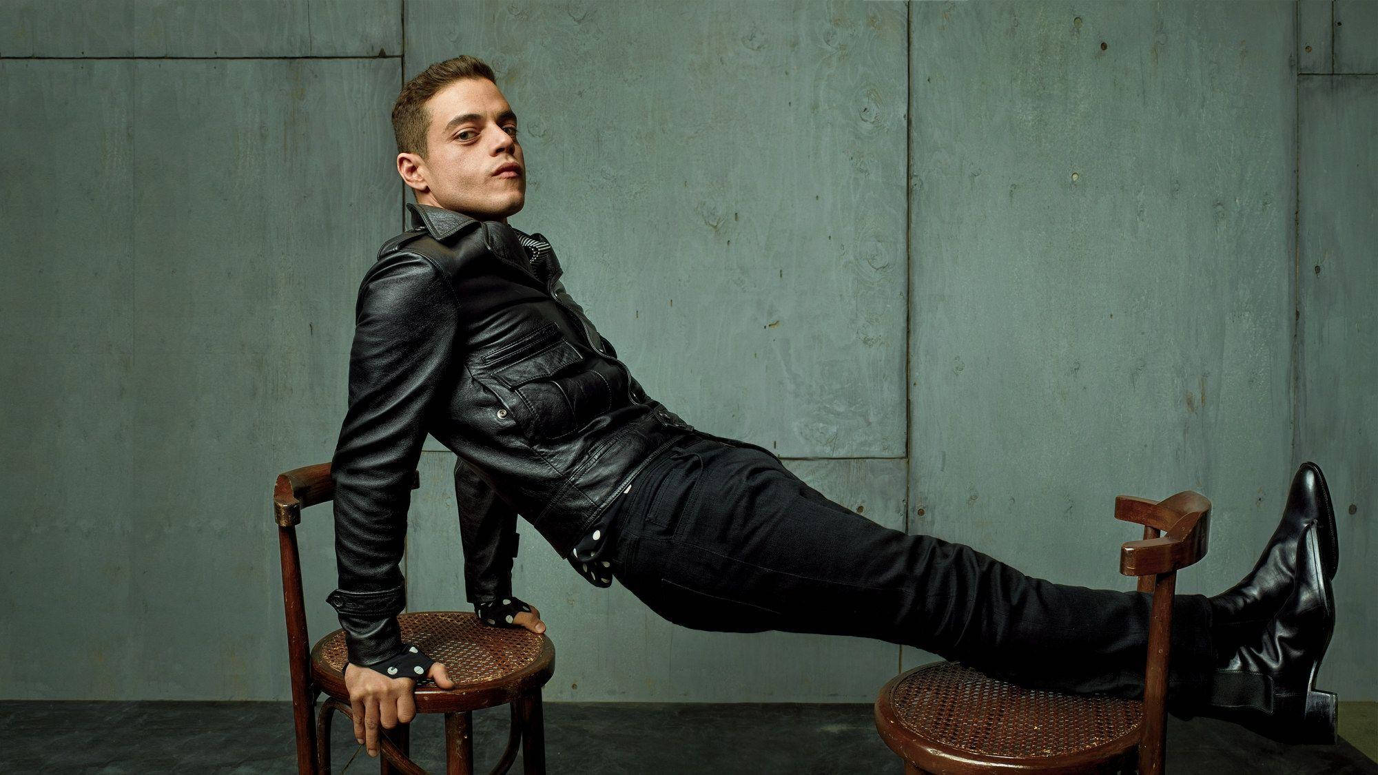 Rami Malek 桌布 壁紙 — 一位身穿黑色皮夾克的年輕男子，自信地靠在兩張木椅上，背景是紋理灰色牆壁。