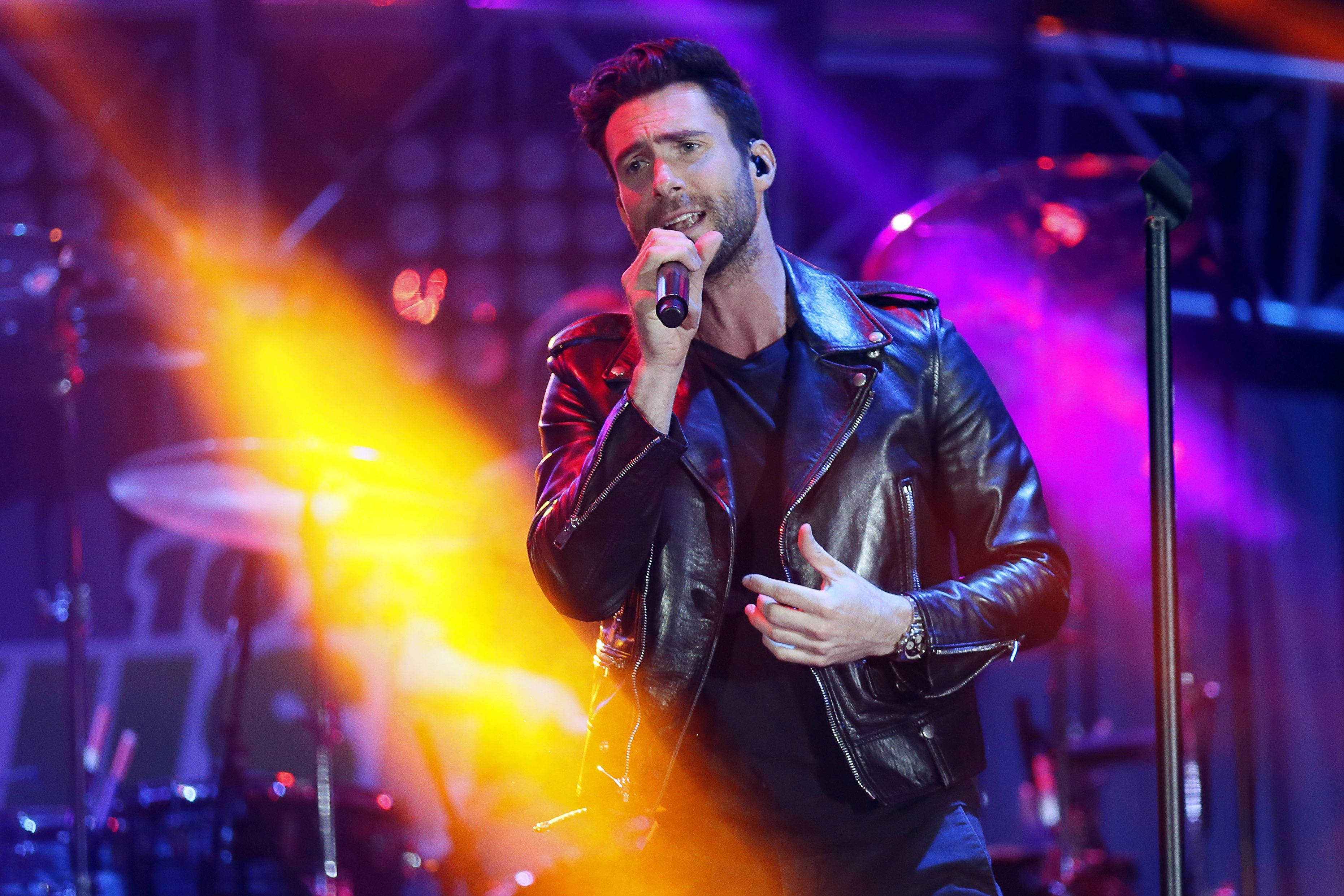 Adam Levine 桌布 壁紙 — 一位身穿黑色皮夾克的男歌手在充滿活力的舞台上，被紫色和橙色燈光照耀下熱情演出。