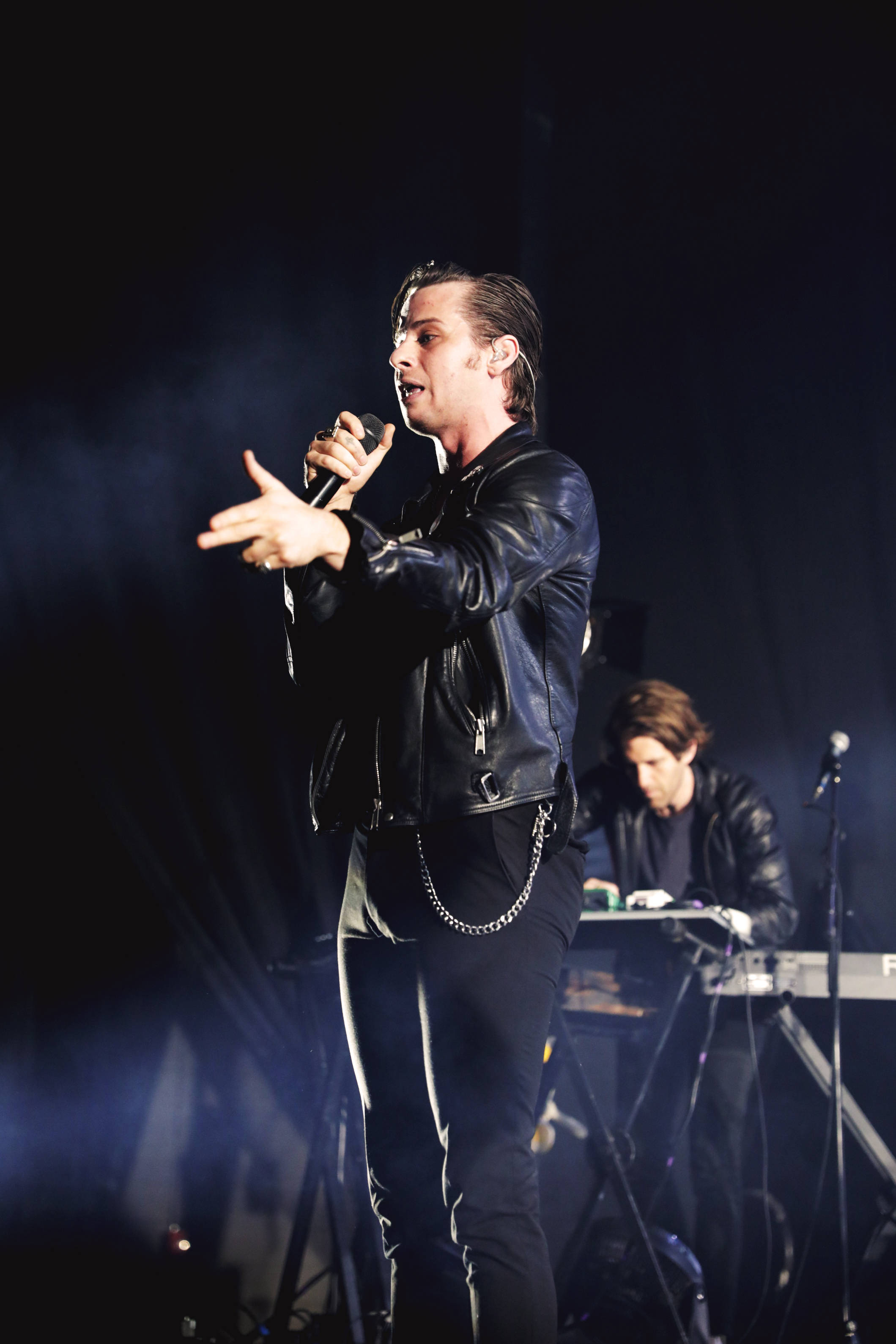 foster the people 桌布 壁紙 — 一位身穿黑色皮夾克的男歌手在燈光昏暗的舞台上充滿激情地表演，背景有煙霧和一位鍵盤手。