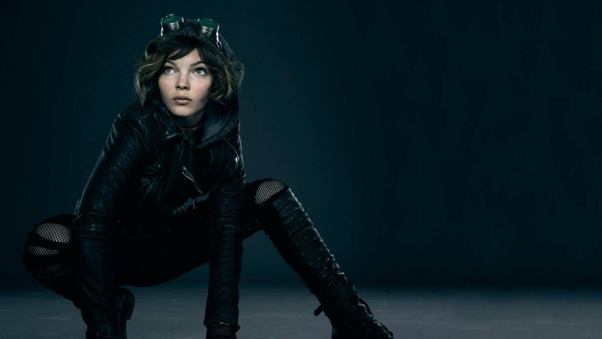 Catwoman 桌布 壁紙 — 一位身穿黑色皮革的年輕女子，頭戴護目鏡，警覺地蹲在深藍色背景前。