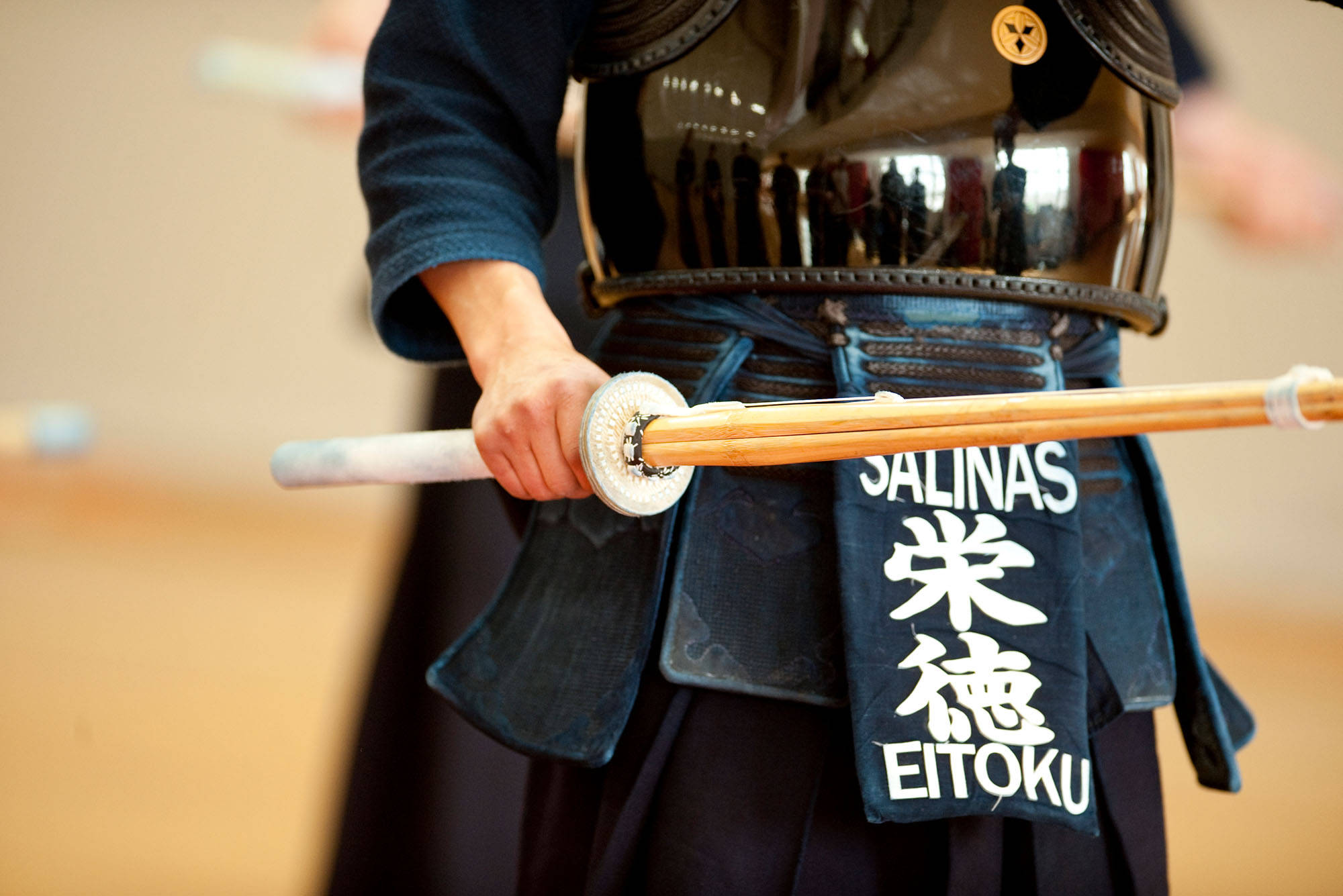 sword 桌布 壁紙 — 一位身穿黑色盔甲的武士，在模糊的室內背景下握著一把白色刀柄的木劍。