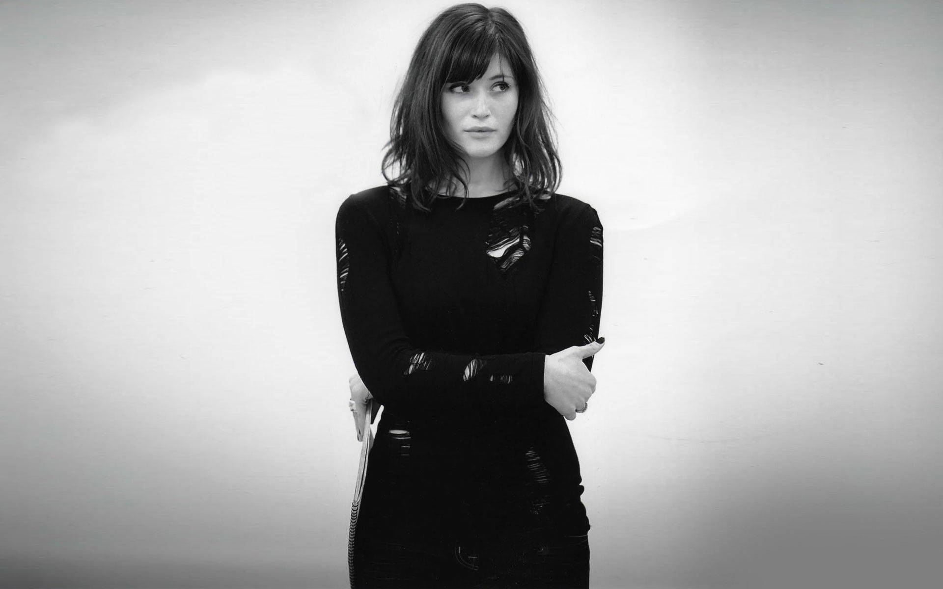Gemma Arterton 桌布 壁紙 — 一位身穿黑色破洞毛衣的女性，在單色灰色背景前交叉雙臂站立。