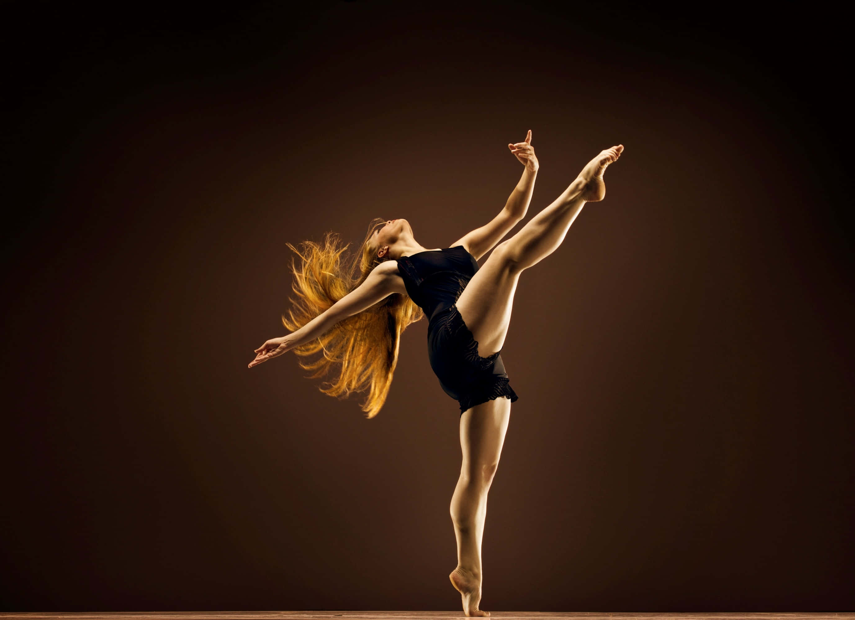 dancer 桌布 壁紙 — 一位身穿黑色緊身衣的優雅舞者，在深色工作室背景下，以飄逸的金色頭髮展現動感姿勢。