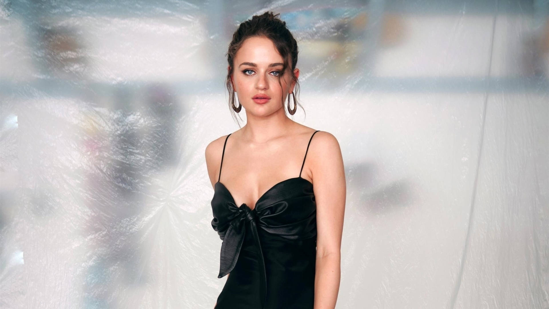 Joey King 桌布 壁紙 — 一位身穿黑色緞面連衣裙、繫著蝴蝶結的女性，自信地站在帶有柔和藍色高光的紋理白色背景前。