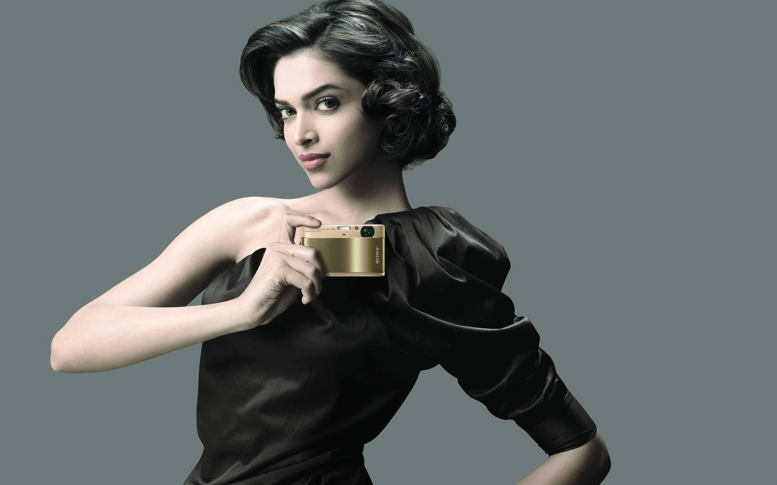 Deepika 桌布 壁紙 — 一位身穿黑色緞面連衣裙的優雅女性，在柔和的灰色背景前手持金色相機。