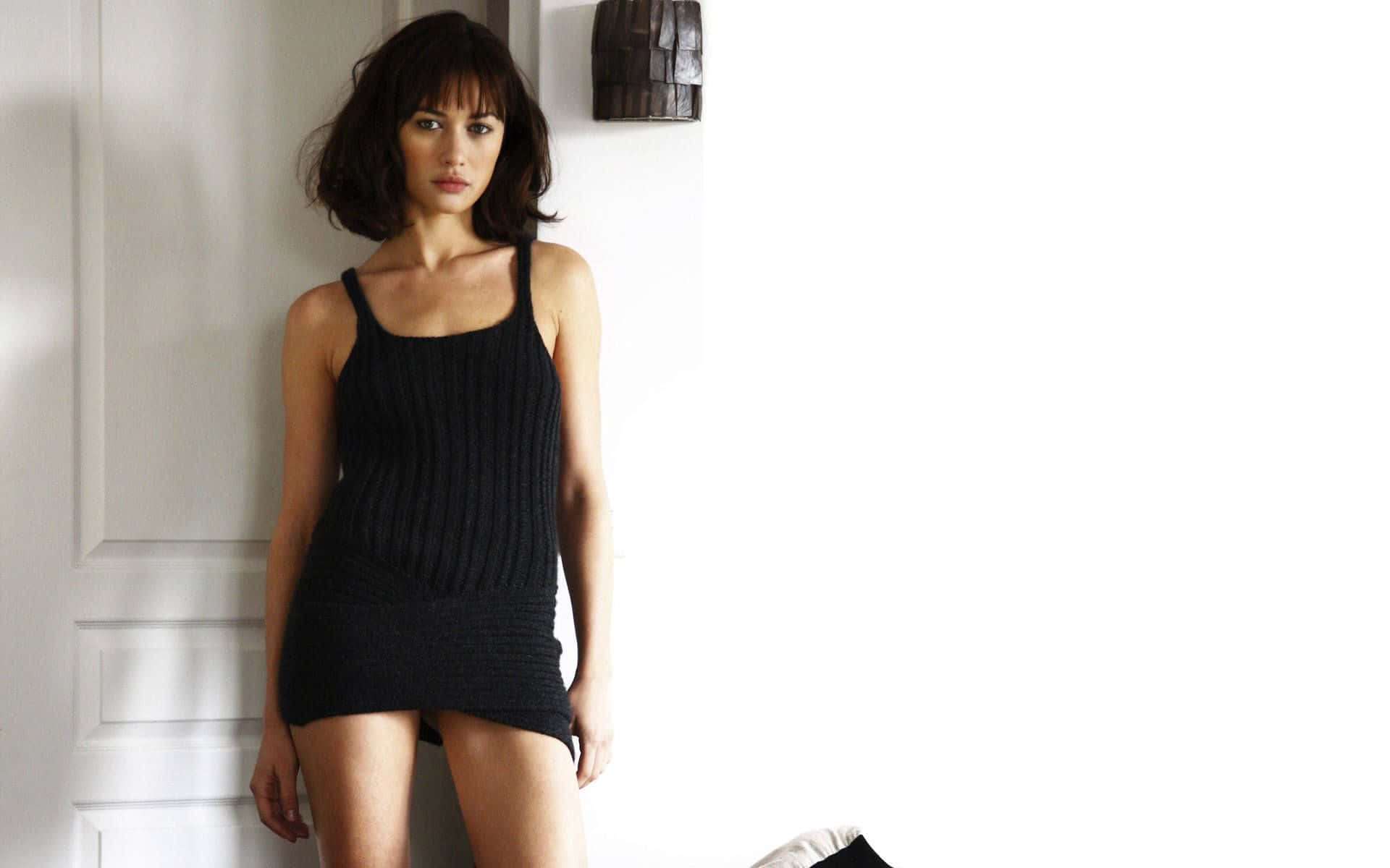 Olga Kurylenko 桌布 壁紙 — 一位身穿黑色羅紋連衣裙的女性站在白色鑲板牆前，柔和自然光投下陰影。
